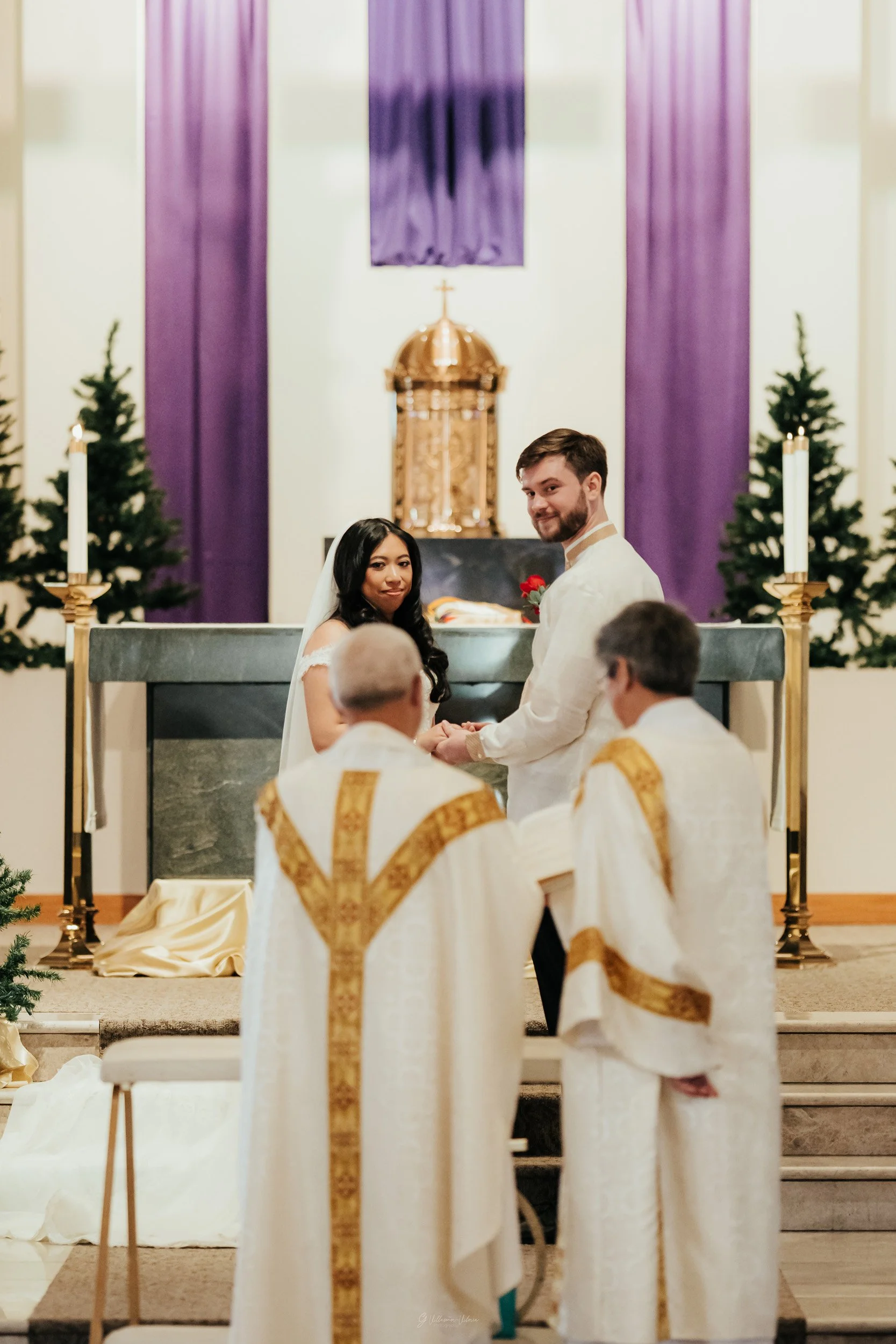 A wedding ceremony taking place at a church altar with a bride and groom holding hands, surrounded by clergy and purple decor.