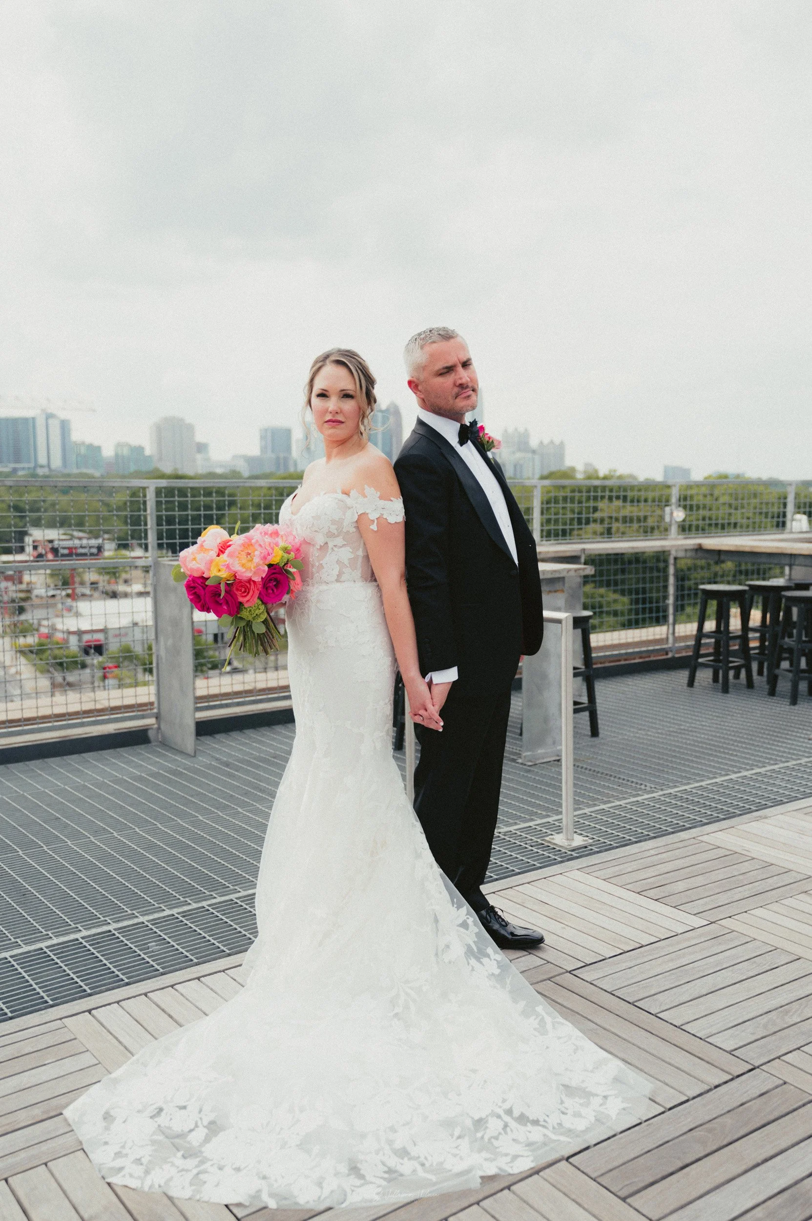 Bride and groom holding hands on a rooftop, bride in white wedding dress with floral lace details holding a colorful bouquet, groom in black tuxedo with bow tie, city skyline in the background.