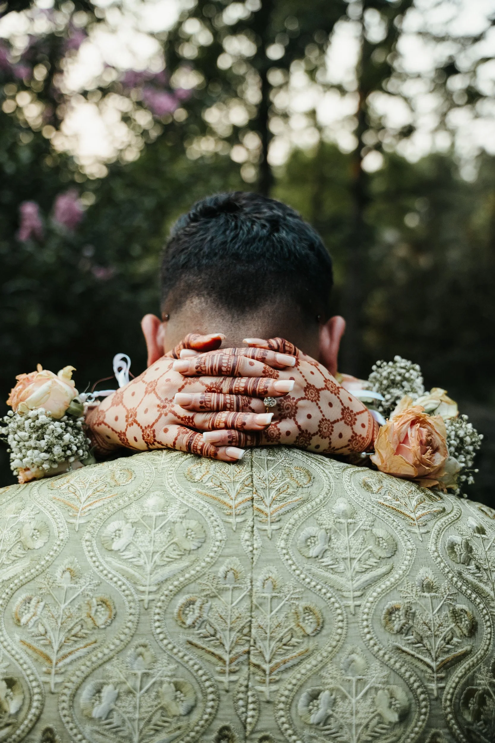 Couple hug outdoors with the woman’s hands with henna designs resting on the man’s shoulders, surrounded by flowers, in a forest setting.