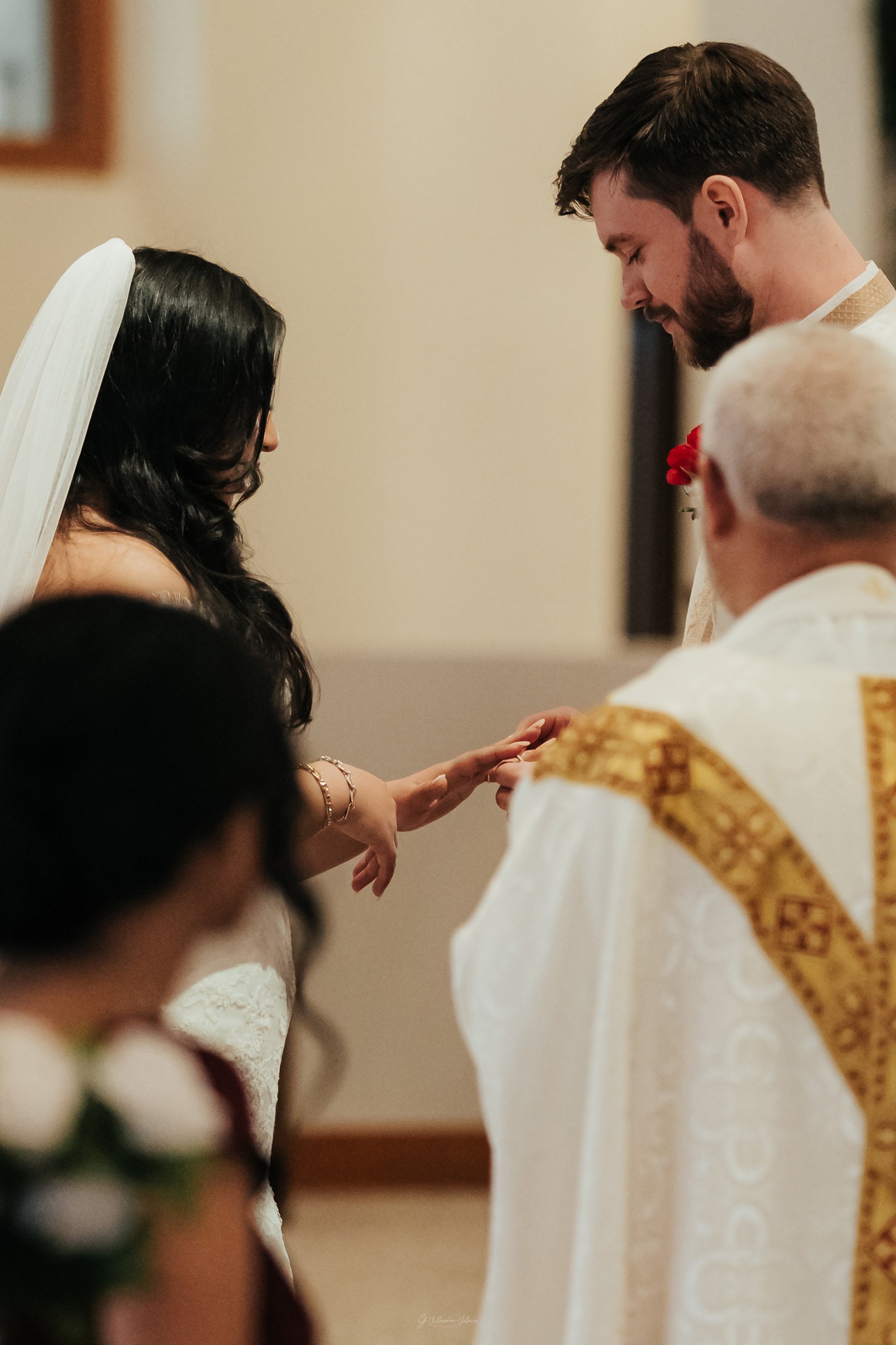A bride and groom exchanging rings during their wedding ceremony, with an officiant present, in an indoor setting.