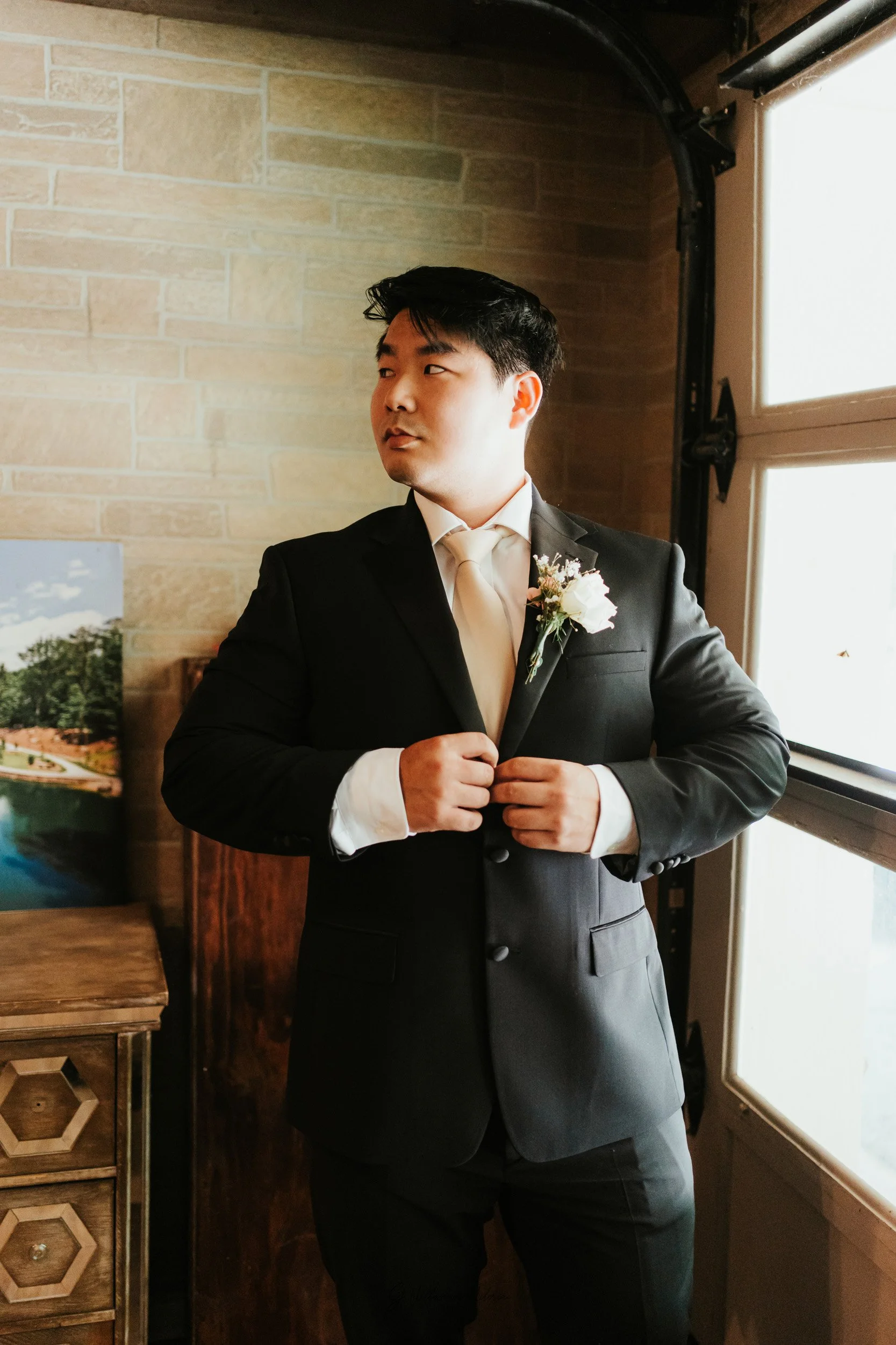 A man in a black suit, white shirt, and white tie adjusting his suit jacket, standing near a window with sunlight coming through, with a boutonniere on his lapel, in a room with a brick wall and wooden furniture.