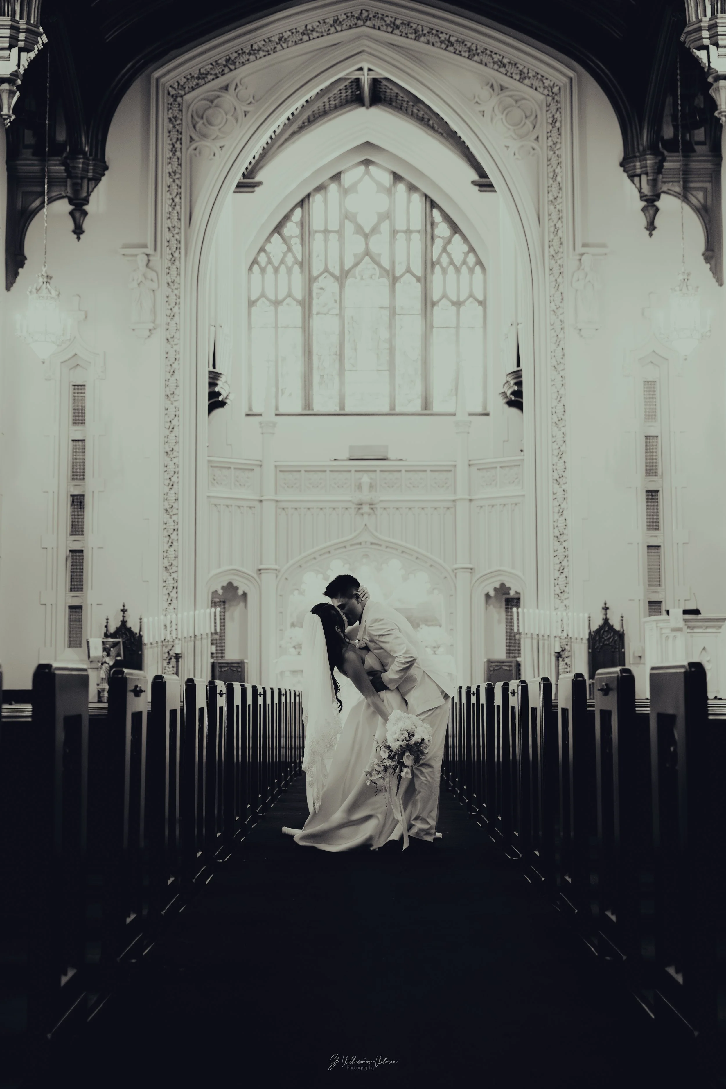 Black and white photo of a bride and groom kissing in the center of a church aisle, with the bride holding a bouquet of flowers.
