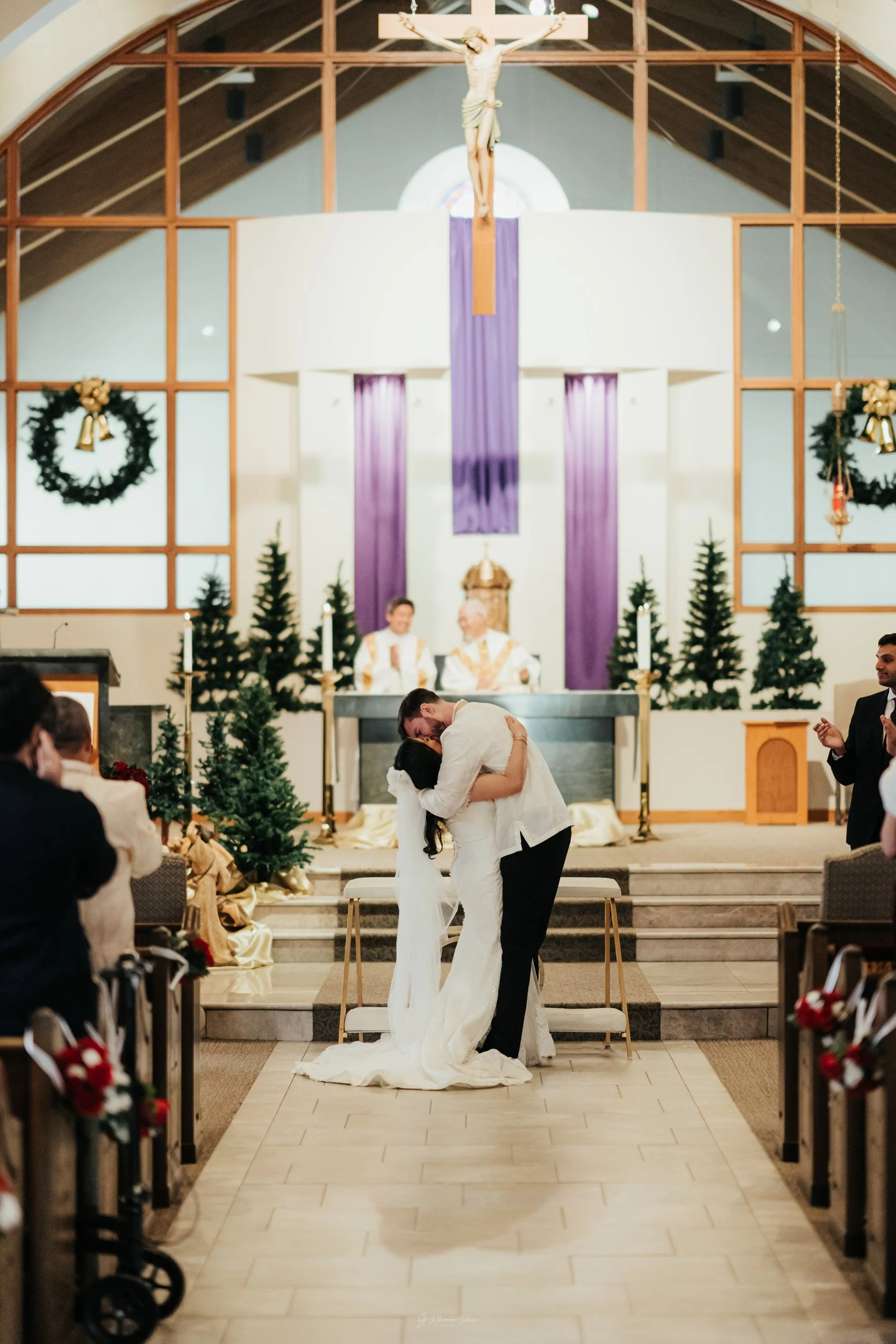 A bride and groom kissing at their wedding ceremony inside a church decorated with Christmas wreaths and small Christmas trees, with priests and guests observing.