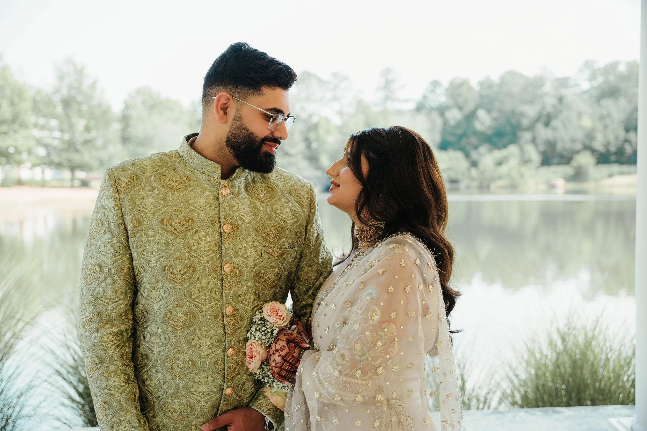 A couple dressed in traditional Indian attire stands close by a lake with trees in the background.