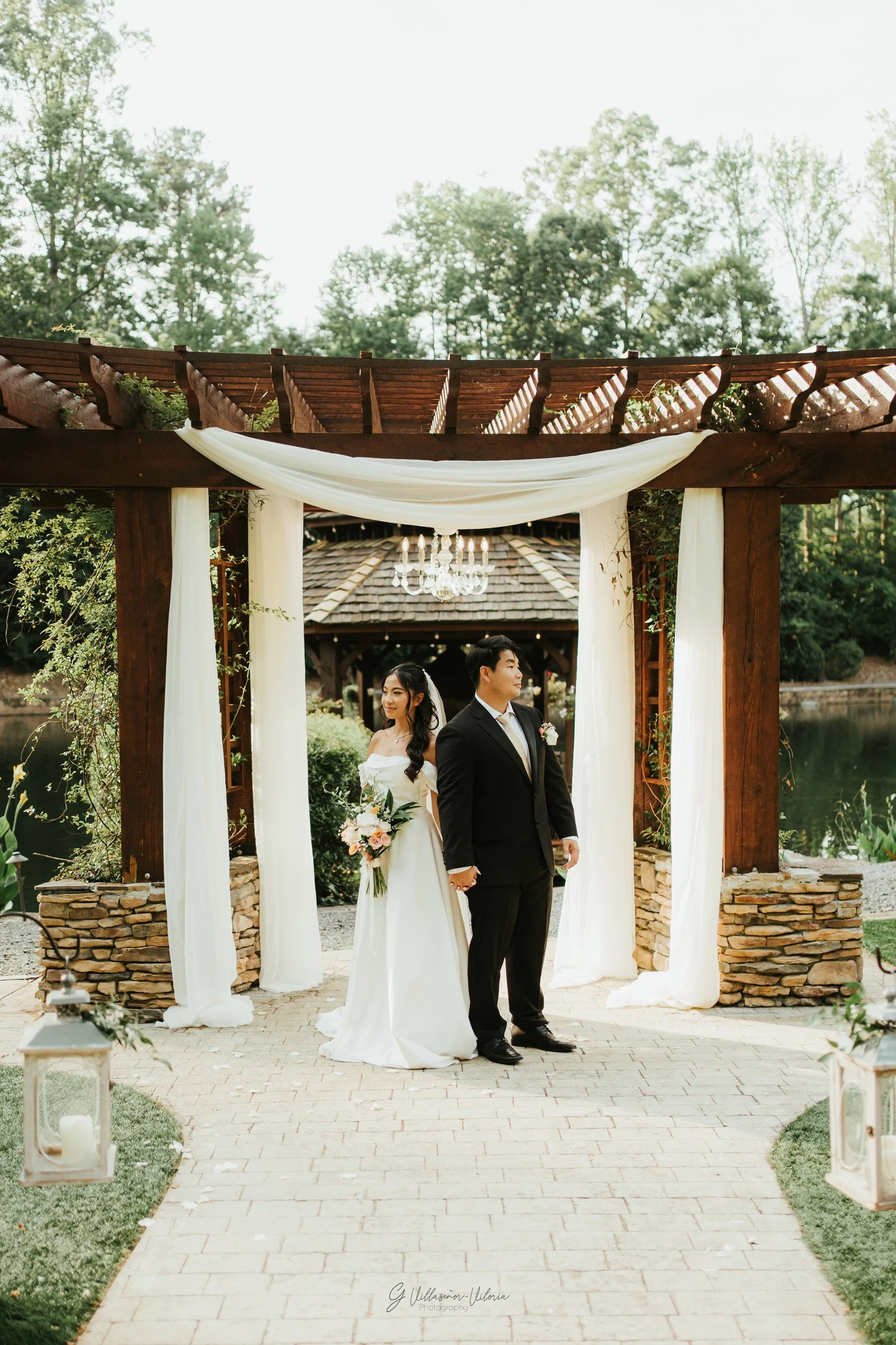 A bride and groom holding hands on their wedding day, standing under a wooden arch with white drapery, near a lake with trees in the background.