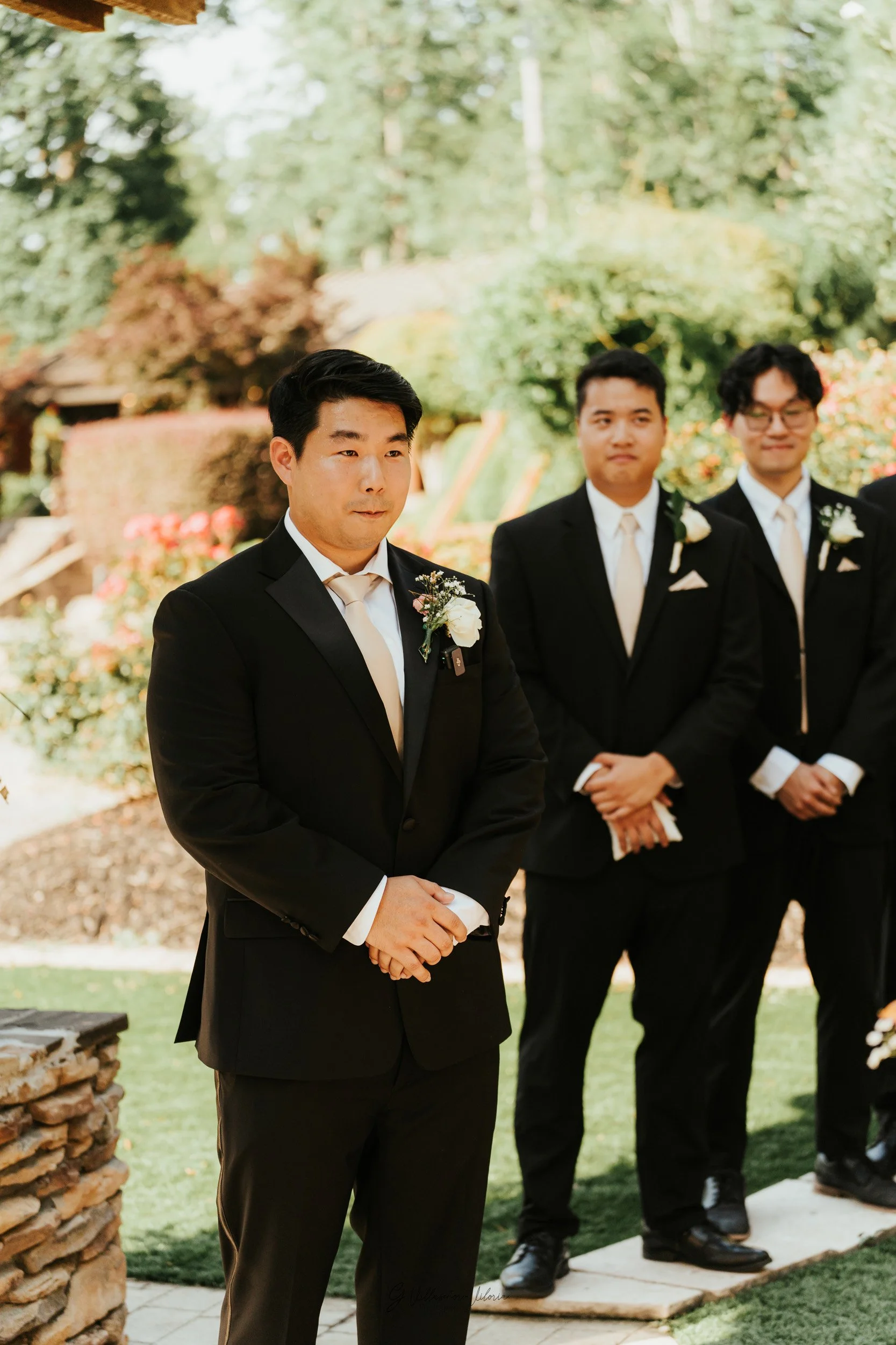 A groom in a black tuxedo with a white tie and boutonniere stands outdoors during a wedding ceremony, with two groomsmen in similar attire standing behind him, in a garden setting with greenery and flowers.