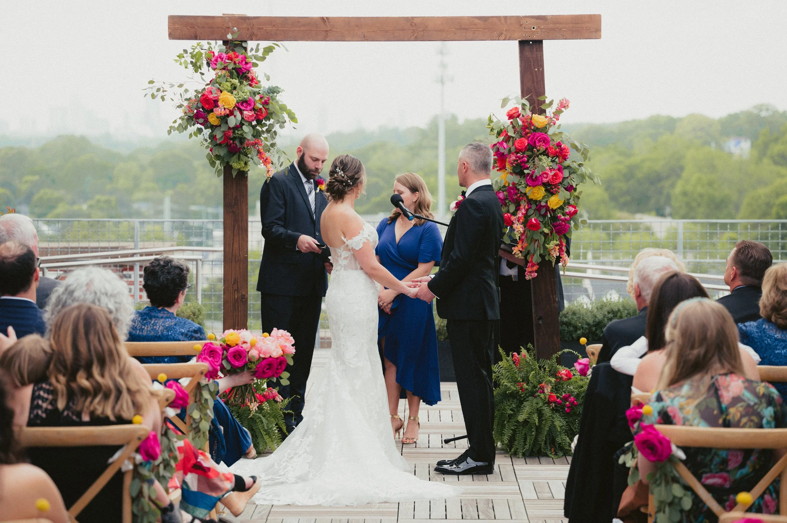 A wedding ceremony taking place outdoors on a wooden deck with a city skyline and green trees in the background. The bride and groom are holding hands and facing each other in front of a wooden arch decorated with colorful flowers. An officiant stand
