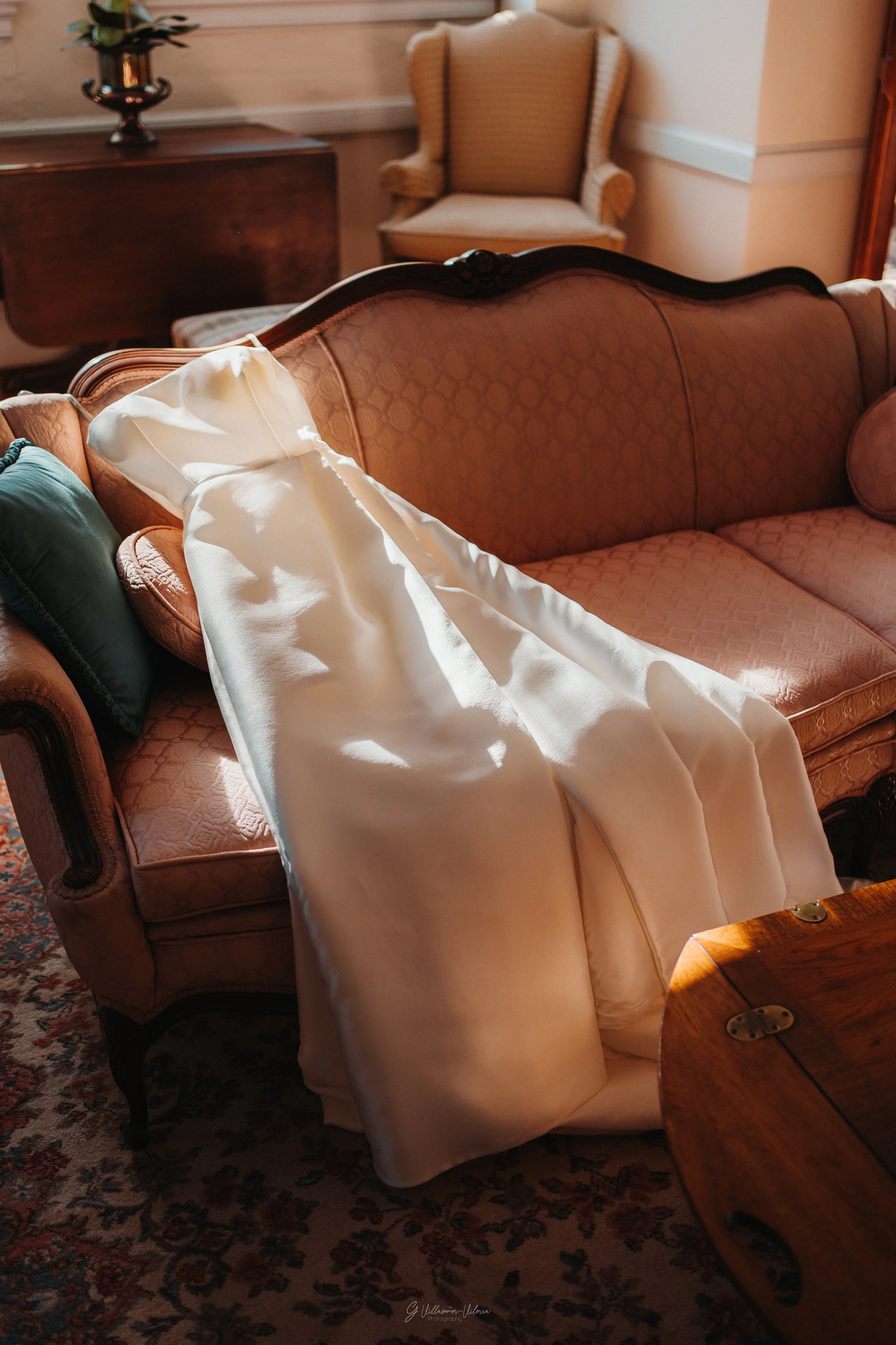 A vintage-style living room with an orange-patterned sofa. A white wedding dress is draped over the sofa's armrest, with sunlight illuminating it. In the background, there is a dark wooden table, a decorative vase, and an armchair with beige upholste
