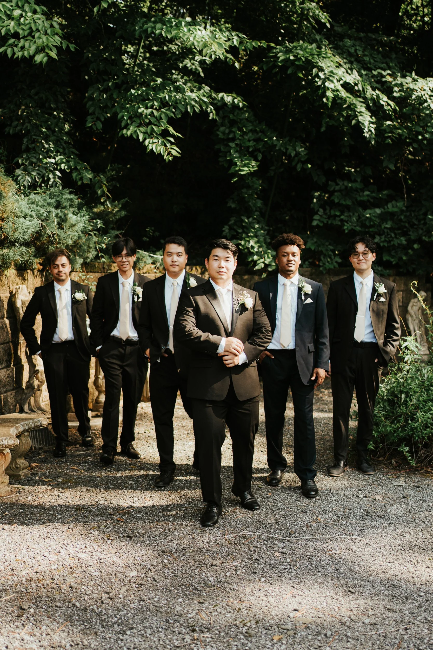 A group of seven men in suits standing outdoors on a gravel path with lush green trees behind them.