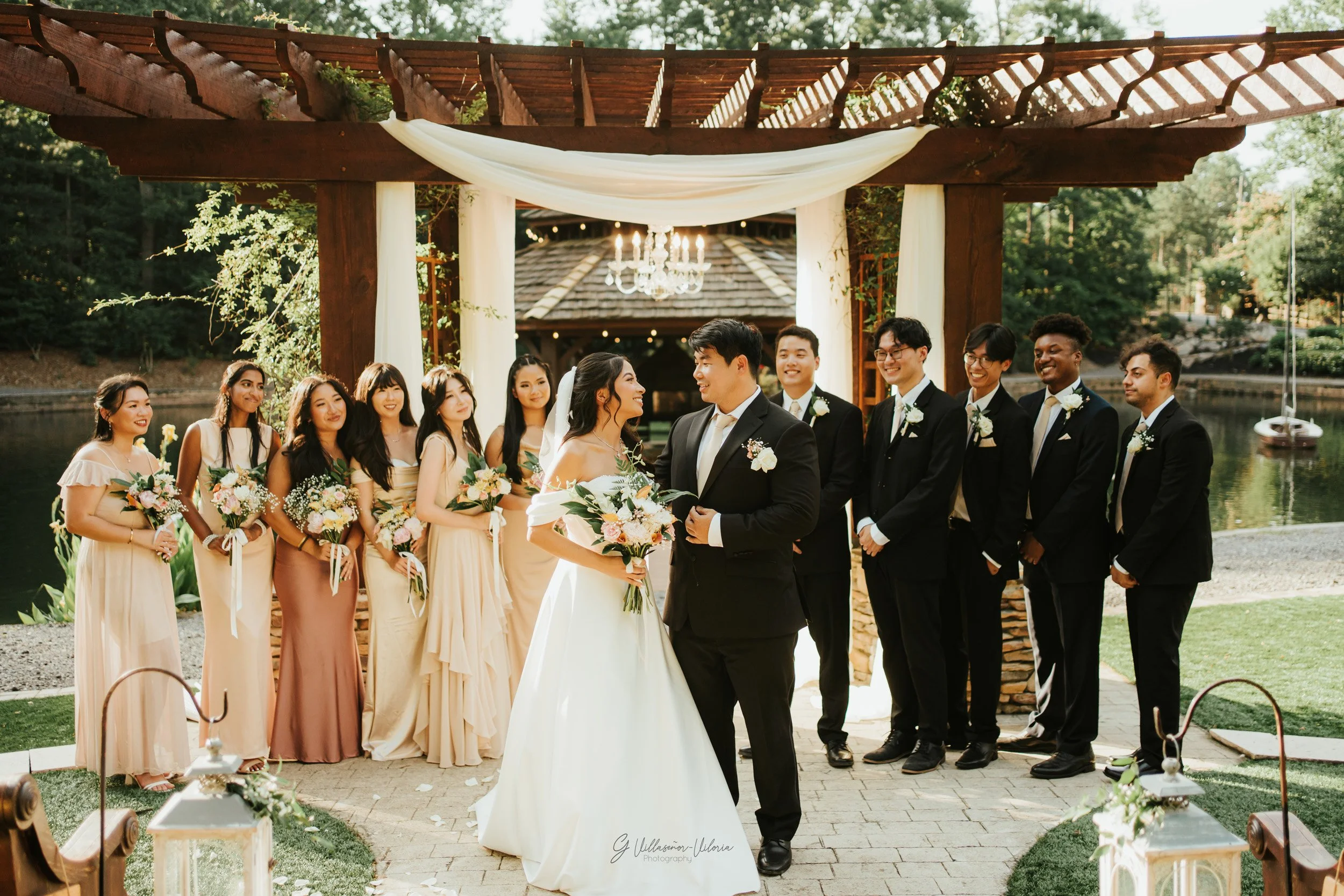 A wedding ceremony outdoors with the bride and groom at the center under a wooden arch draped with white fabric and a chandelier hanging above. The bride is in a white gown holding a bouquet of flowers, and the groom is in a black suit. Bridesmaids a