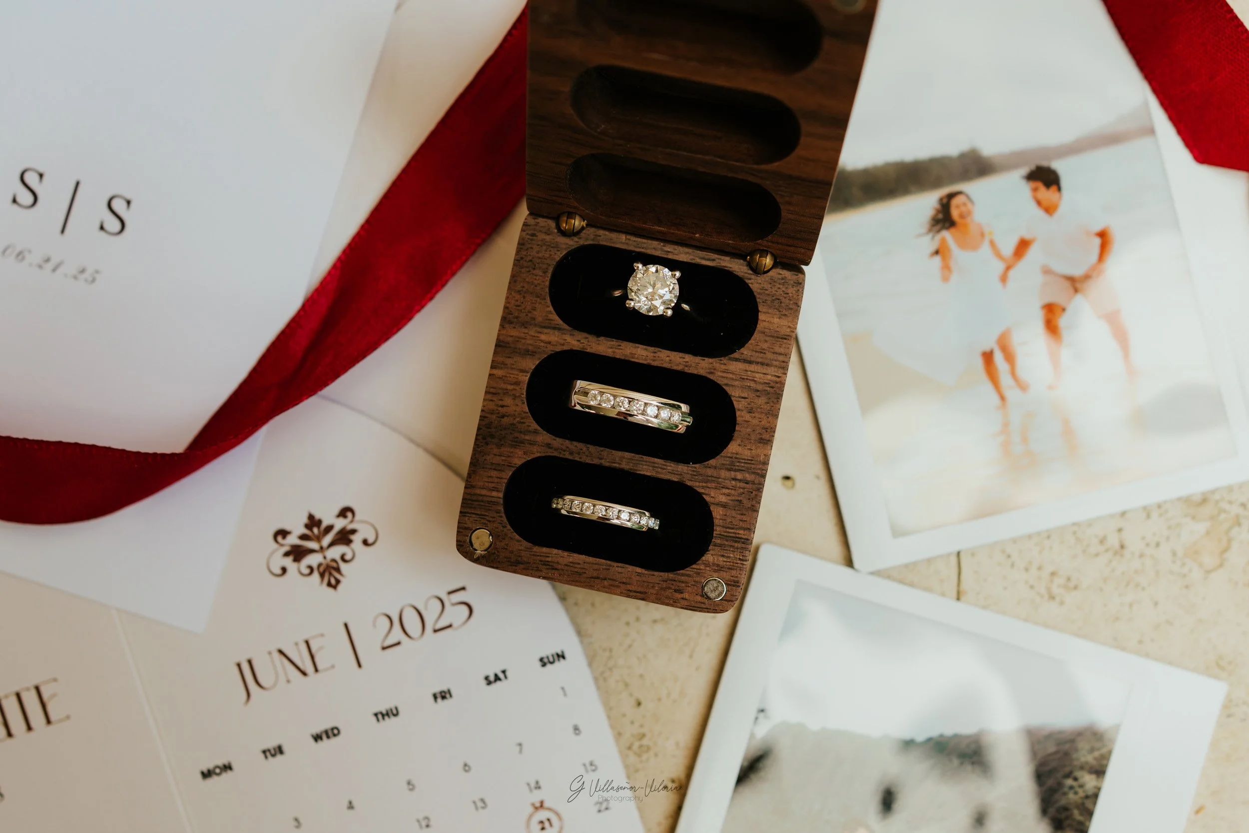 A wooden jewelry box containing a diamond ring and two wedding bands, surrounded by a calendar, photographs, and a white envelope with a red ribbon.