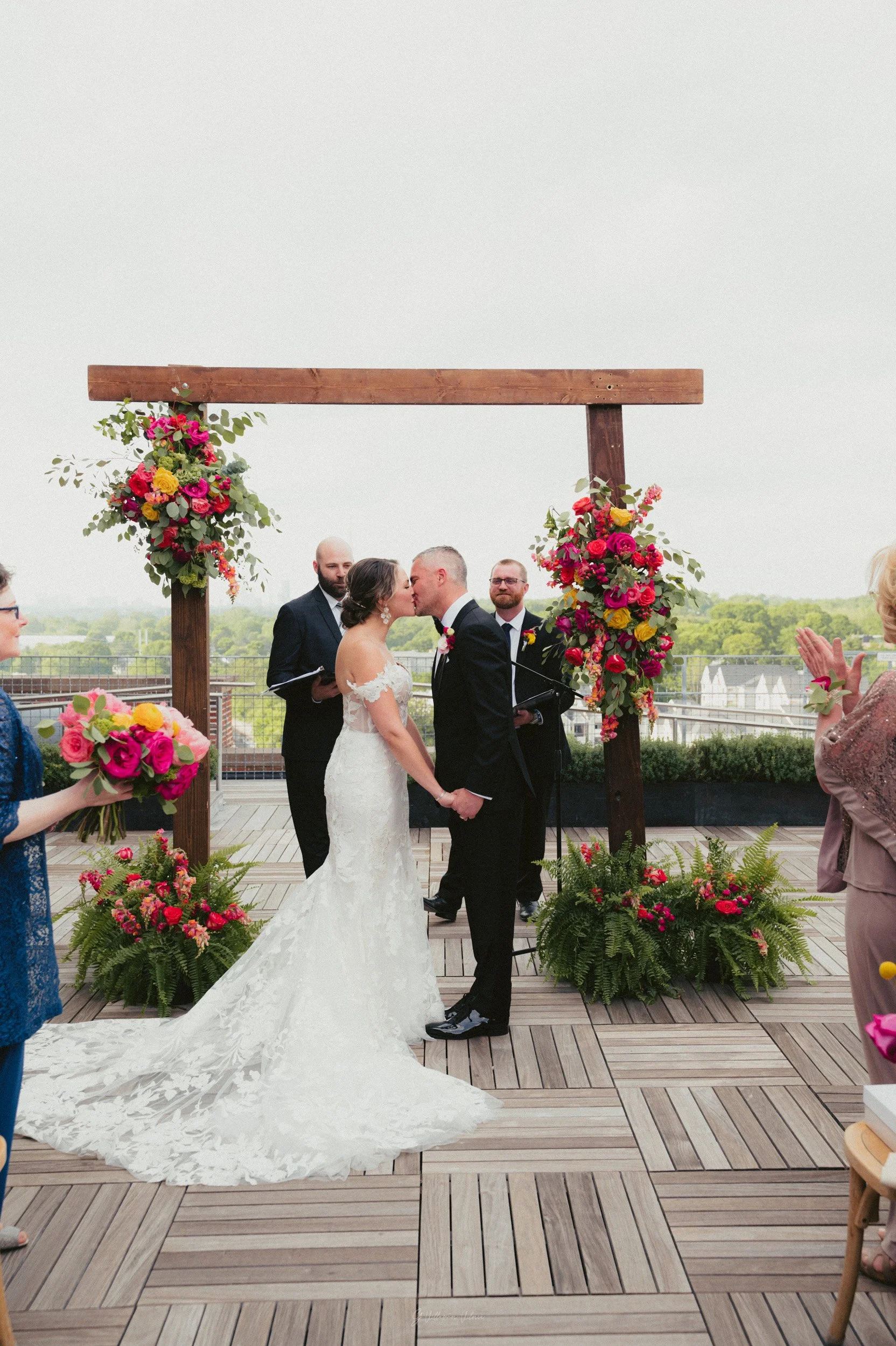 A couple in wedding attire, holding hands and kissing under a wooden arch decorated with colorful flowers, during their wedding ceremony on a rooftop with cityscape in the background.