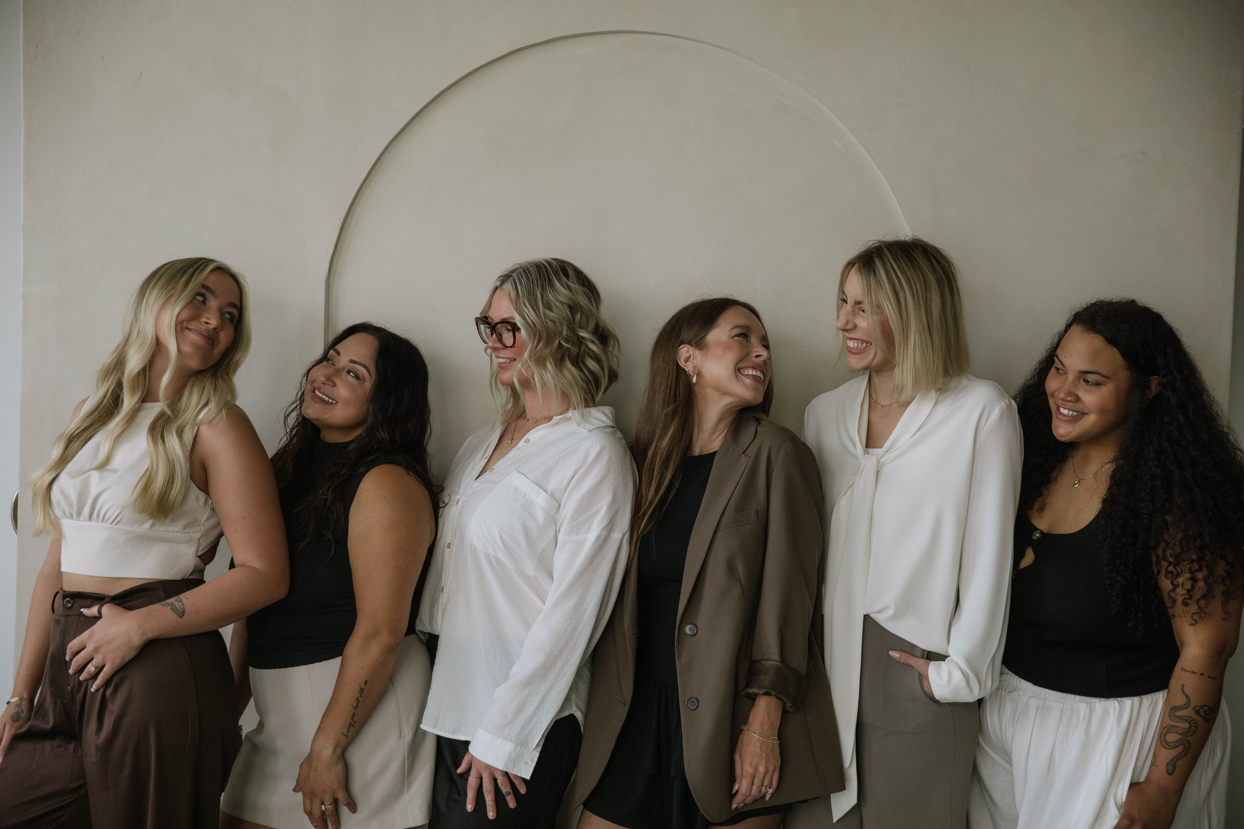 Six women standing side by side against a plain wall, smiling and looking at each other in a friendly and joyful manner.