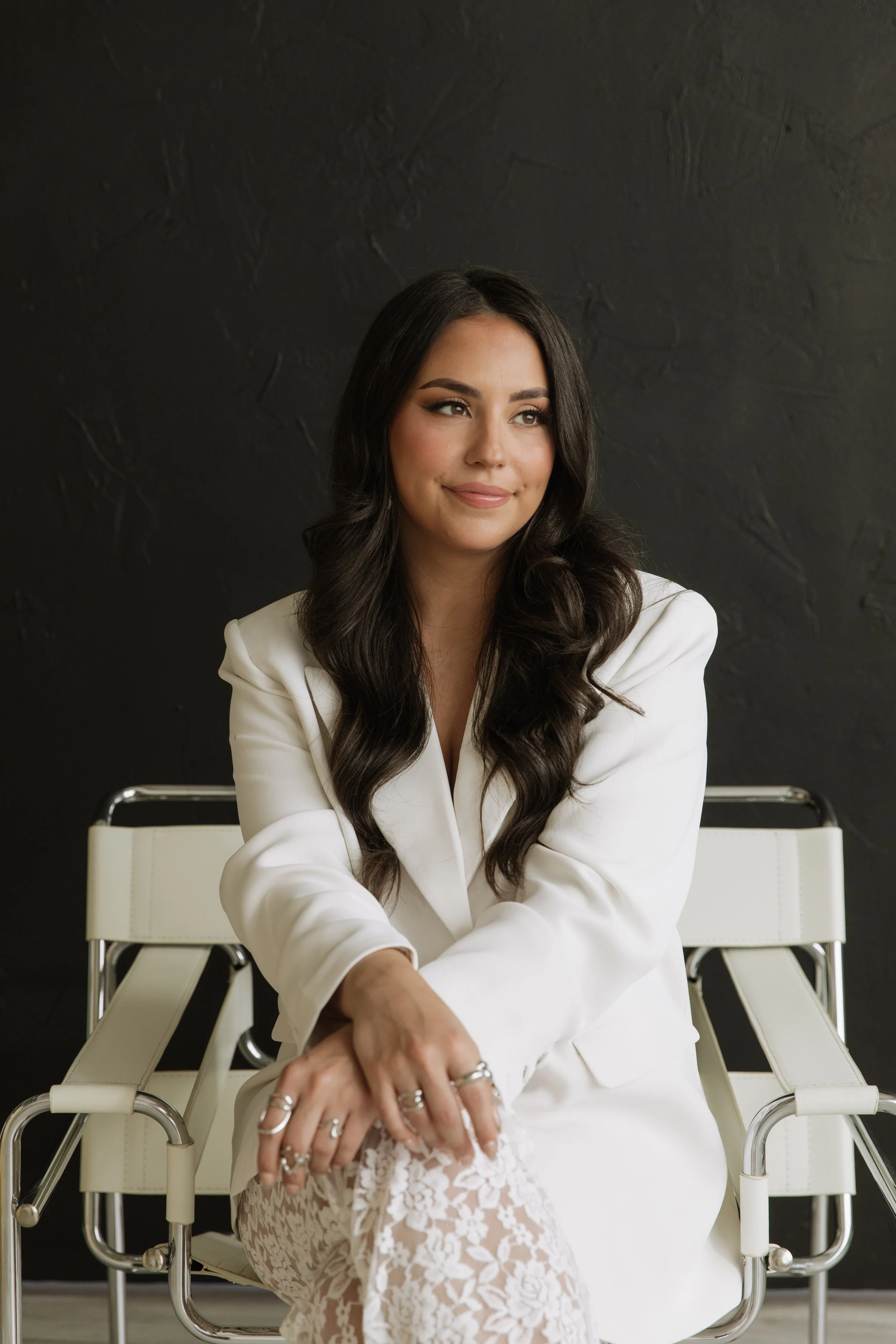 Young woman with long dark wavy hair, wearing a white blazer and lace pants, sitting on a modern white chair with metal accents, against a dark textured wall, smiling softly.