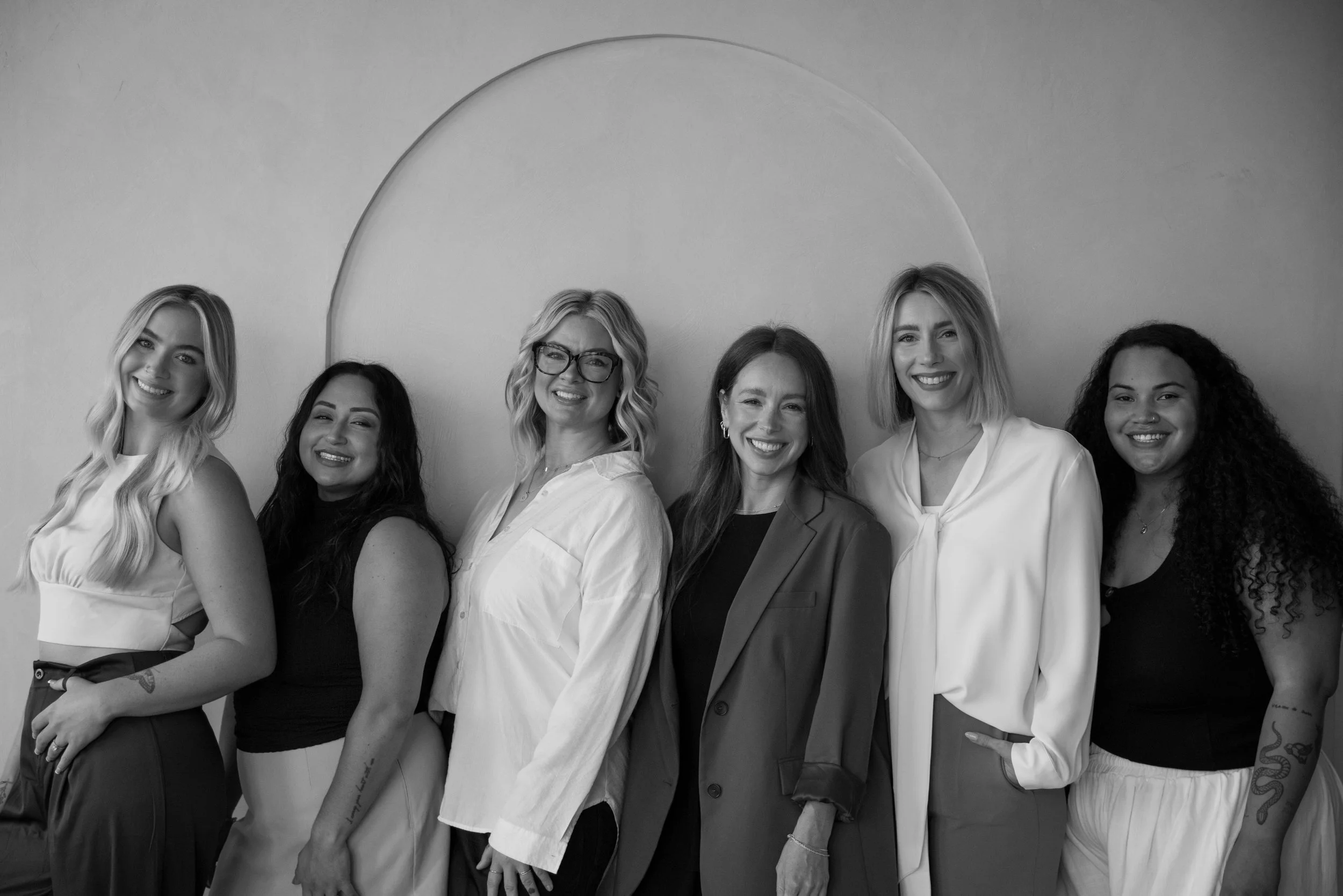 A group of six women standing side by side and smiling in front of a plain wall with a partial circular design.