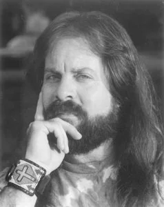 Black and white photo of a person with long hair and a beard, touching their chin, wearing a bracelet.