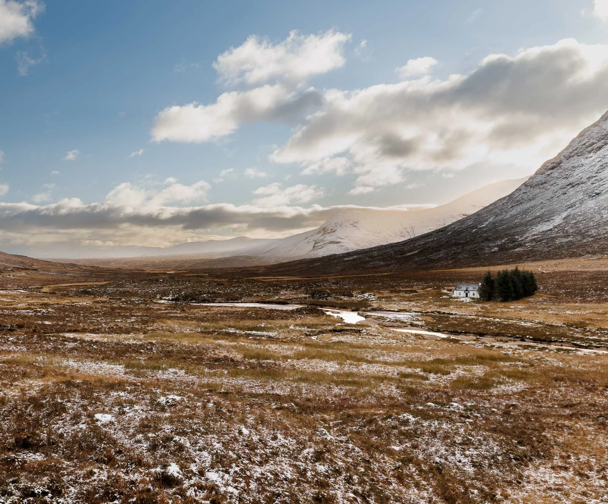 Glencoe, Scotland,  remote landscape featuring snow-capped mountains, a cloudy sky, a grassy plain with patches of snow, a winding stream, and a white house surrounded by trees.