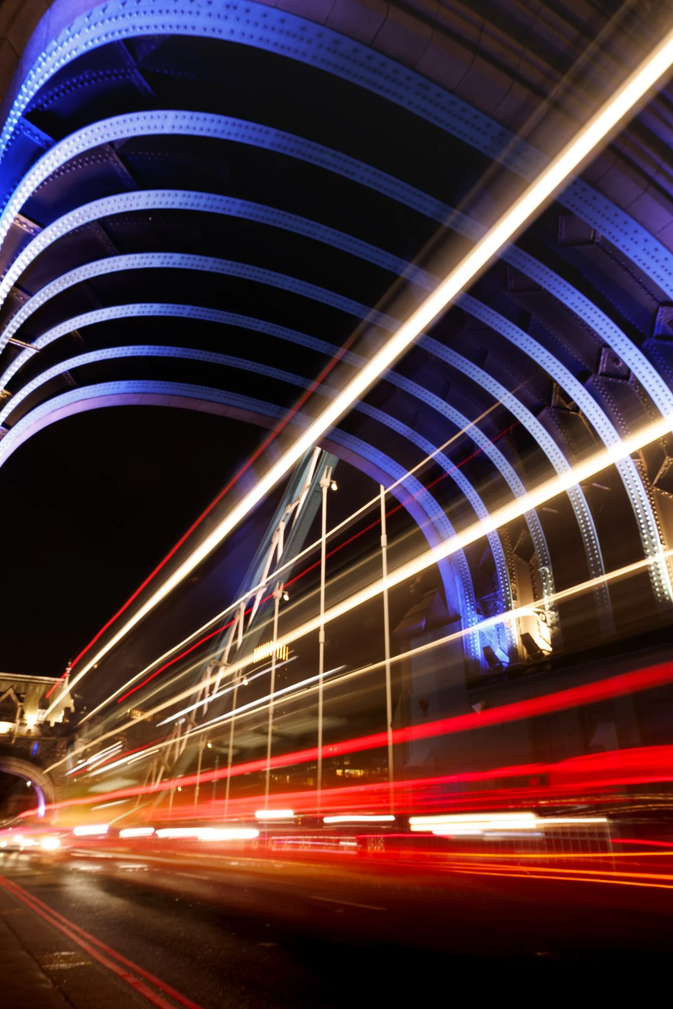 Nighttime Tower Bridge, London, city scene with a brightly lit bridge and moving cars creating light trails, under a large illuminated arch.