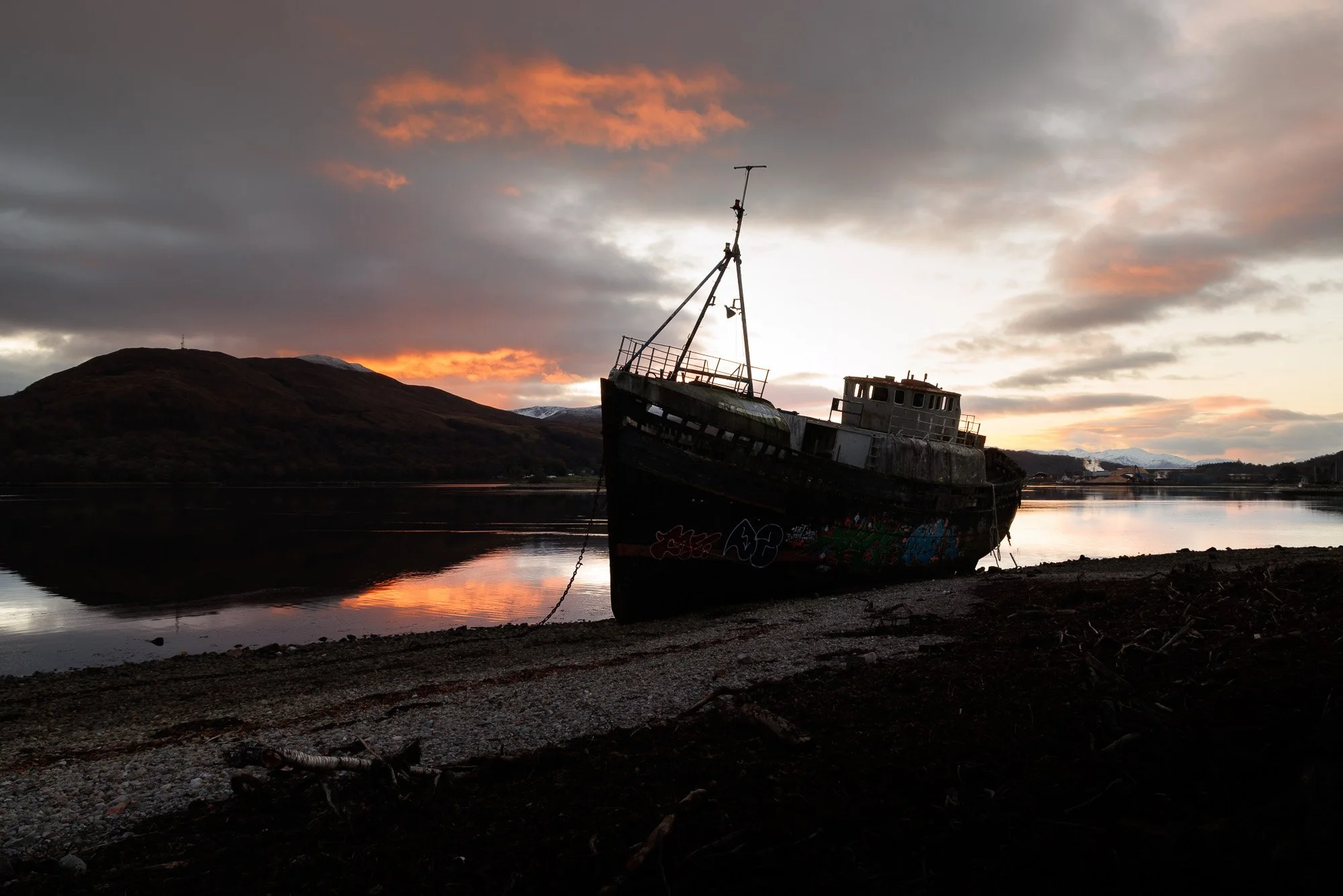 A derelict boat resting on the shore of a calm body of water during sunset, with hills and a partly cloudy sky in the background.