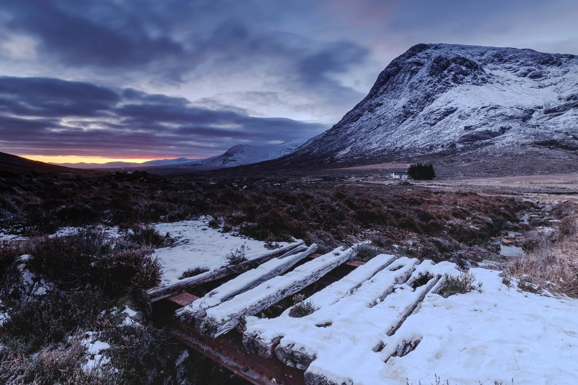glencoe, scotland, with Snow-covered landscape with a large mountain and cloudy sky at sunset, with a small house and trees in the distance and a wooden bench in the foreground.
