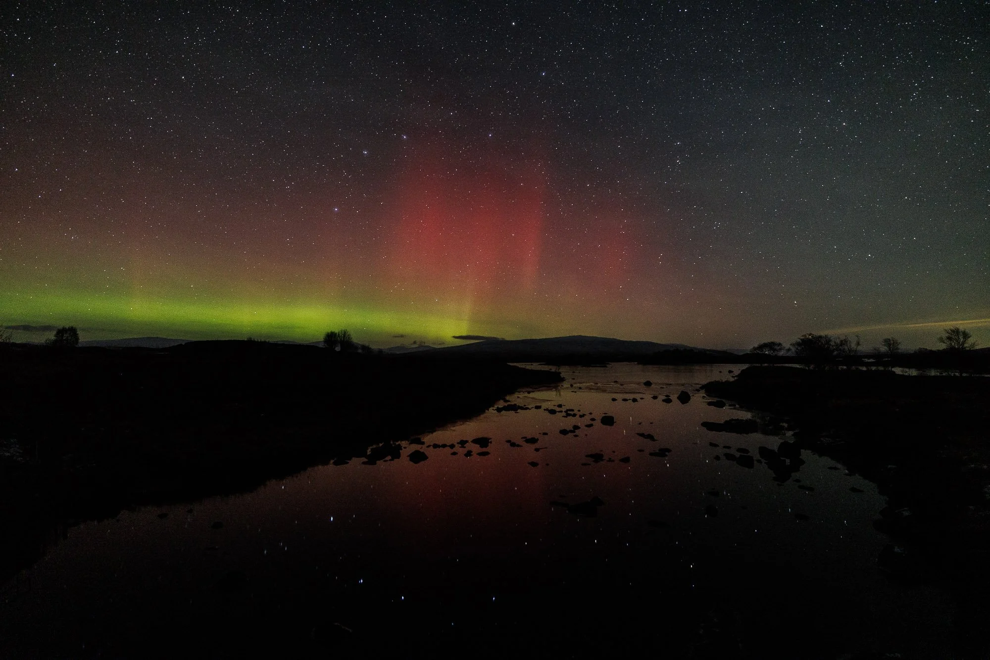 The image shows a Glencoe, Scotland,  sky with numerous stars and the Northern Lights (aurora borealis) displaying green and red colors over a dark landscape with a river and silhouetted trees.