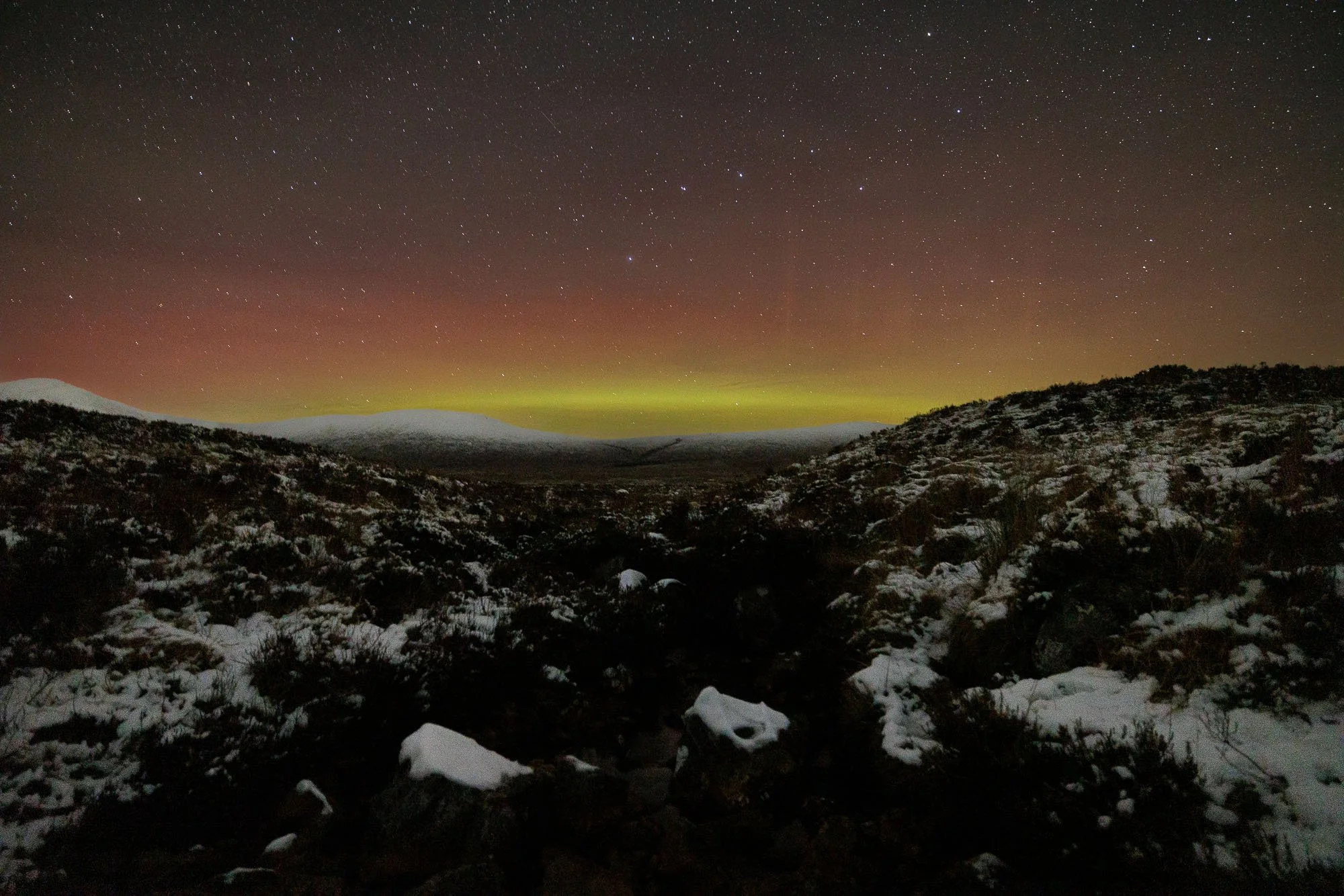 Nighttime landscape with snow-covered ground, mountain silhouettes, and the northern lights in green and red hues across a starry sky.
