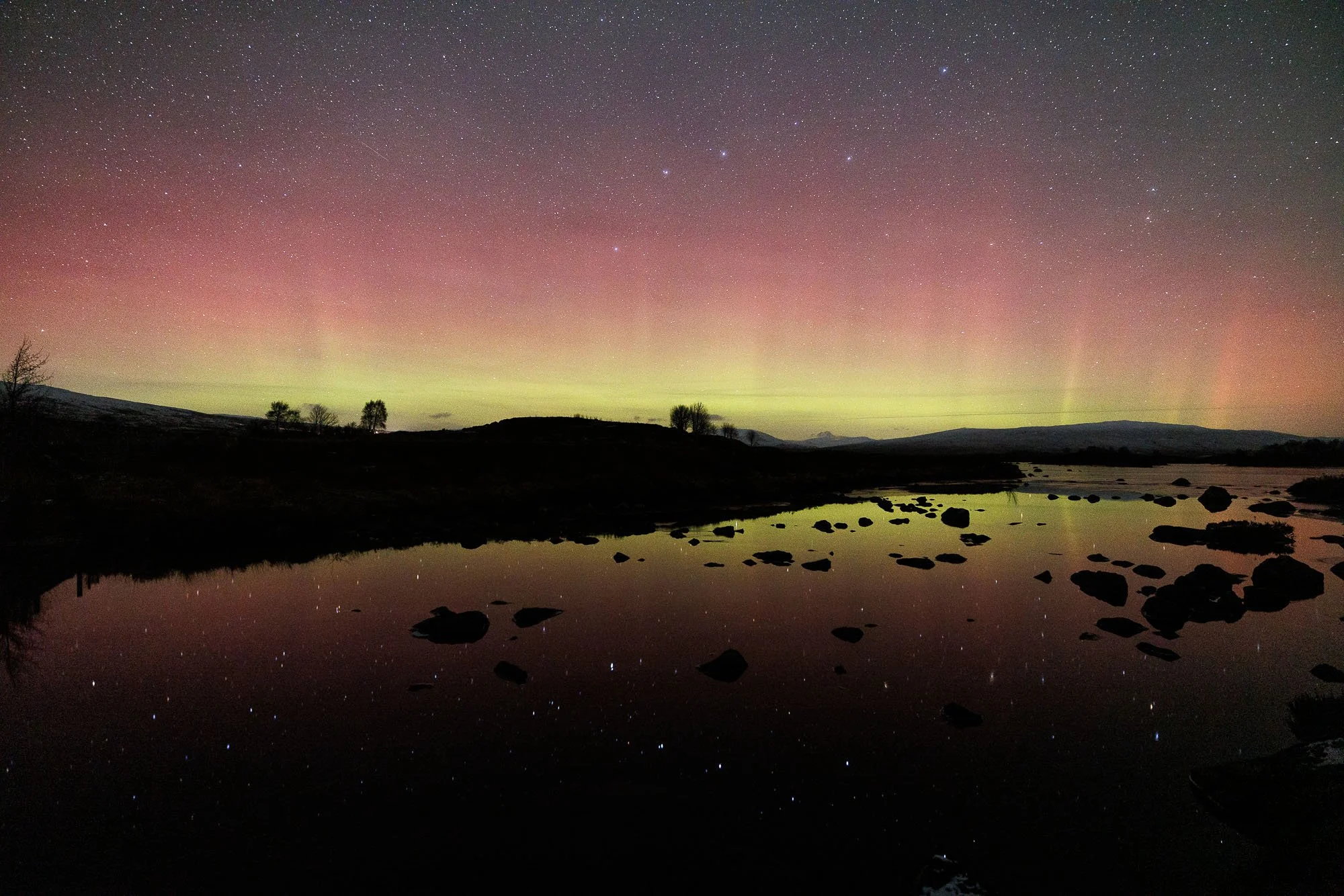 Glencoe, Scotland,  landscape showing a river with rocks, silhouetted trees and hills, and the sky displaying colorful aurora borealis and numerous stars.