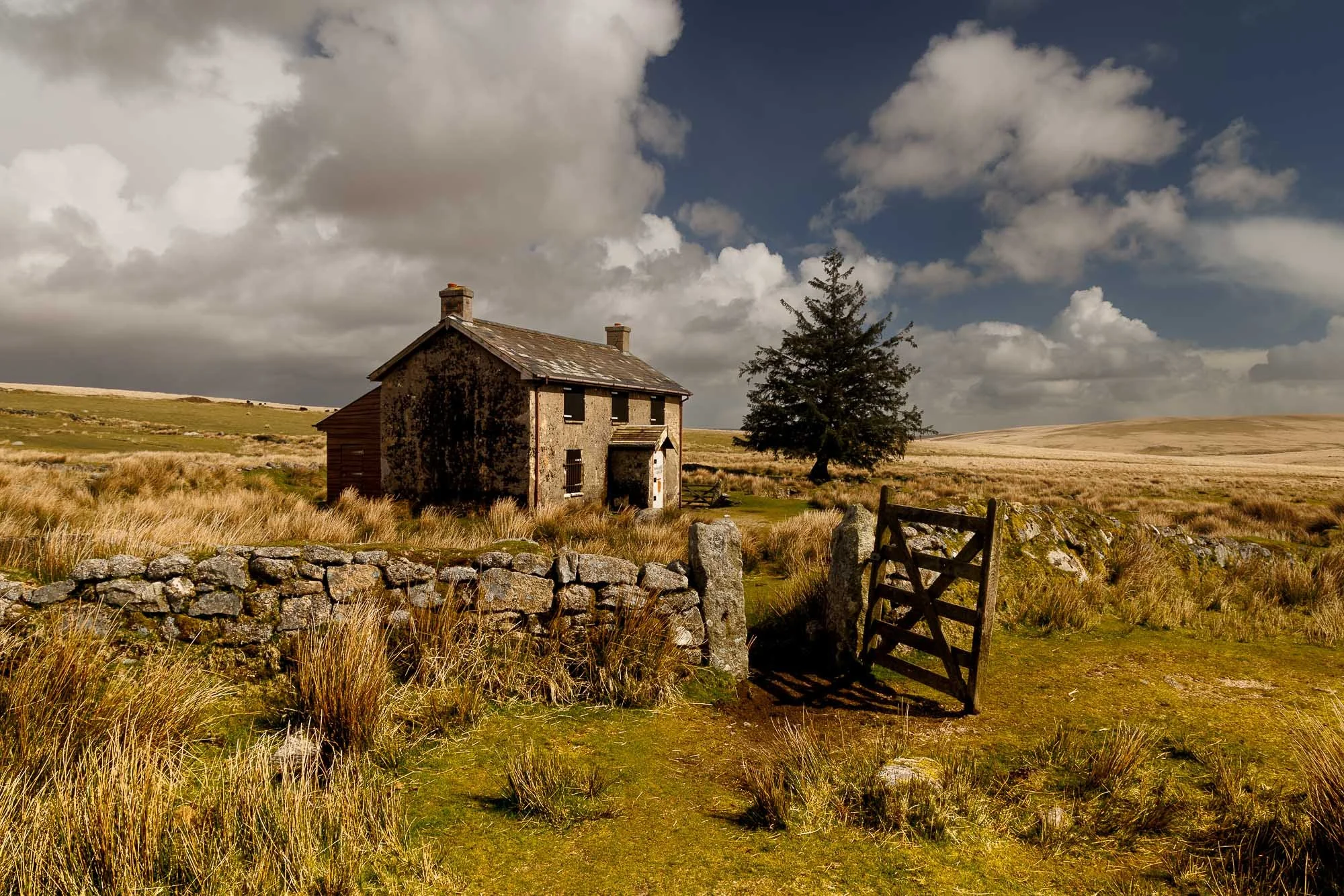 Old stone house surrounded by grassy fields, stone fence, wooden gate, and a large tree under a partly cloudy sky.