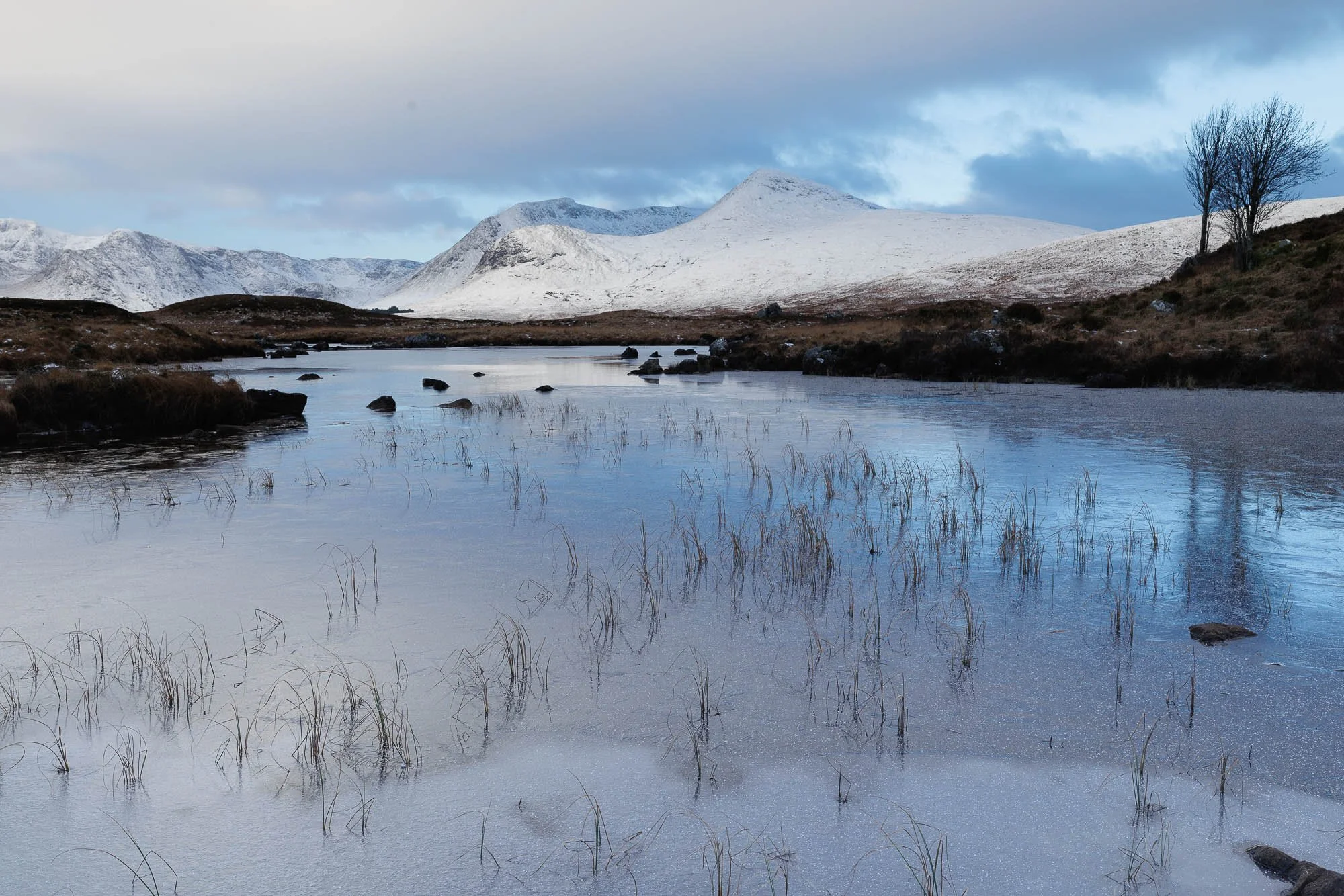 Glencoe, Scotland, snow capped mountains over a calm river with some rocks and sparse grass, under a partly cloudy sky.