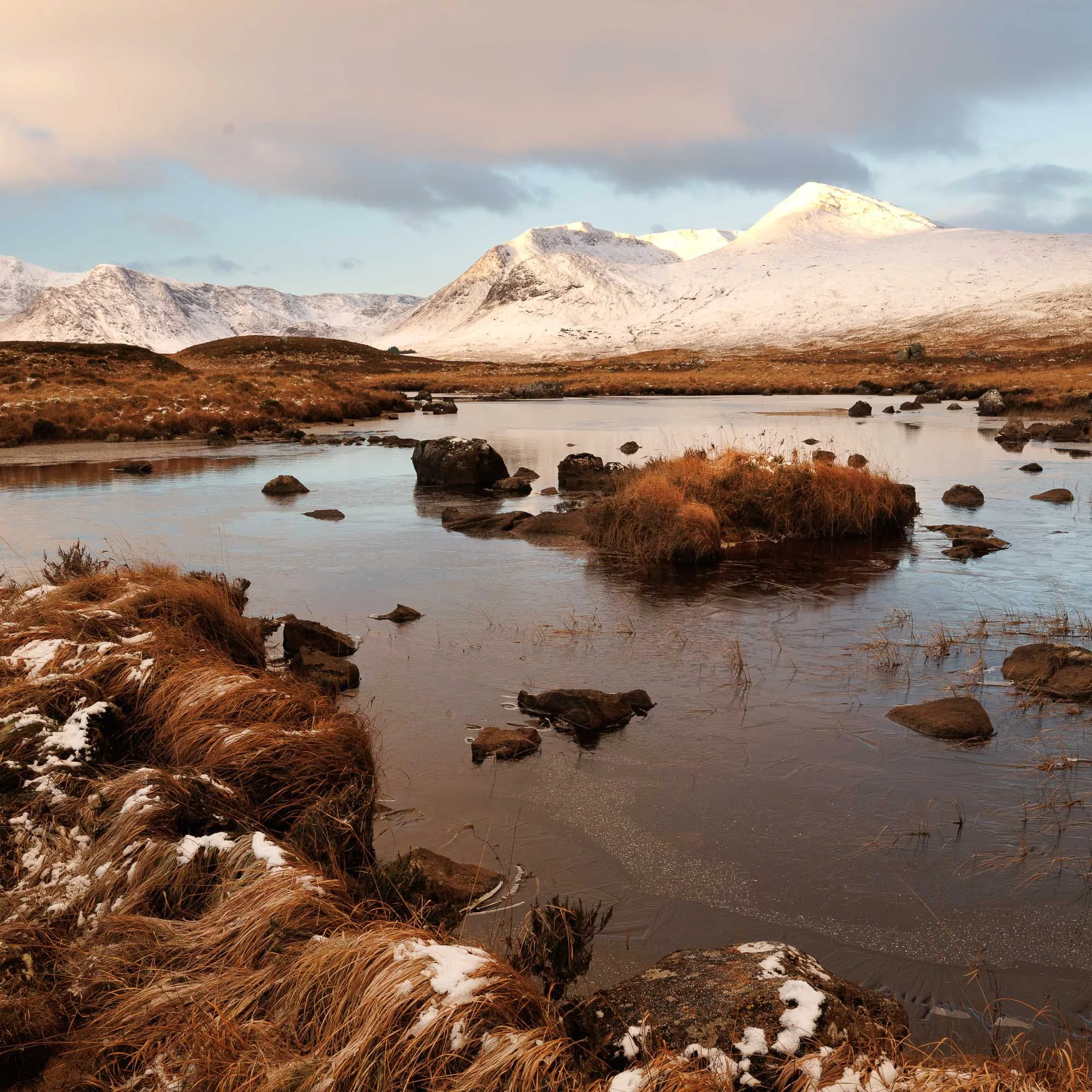 Glencoe, Scotland, snow capped mountains over a calm river with rocks and grassy patches in the foreground.