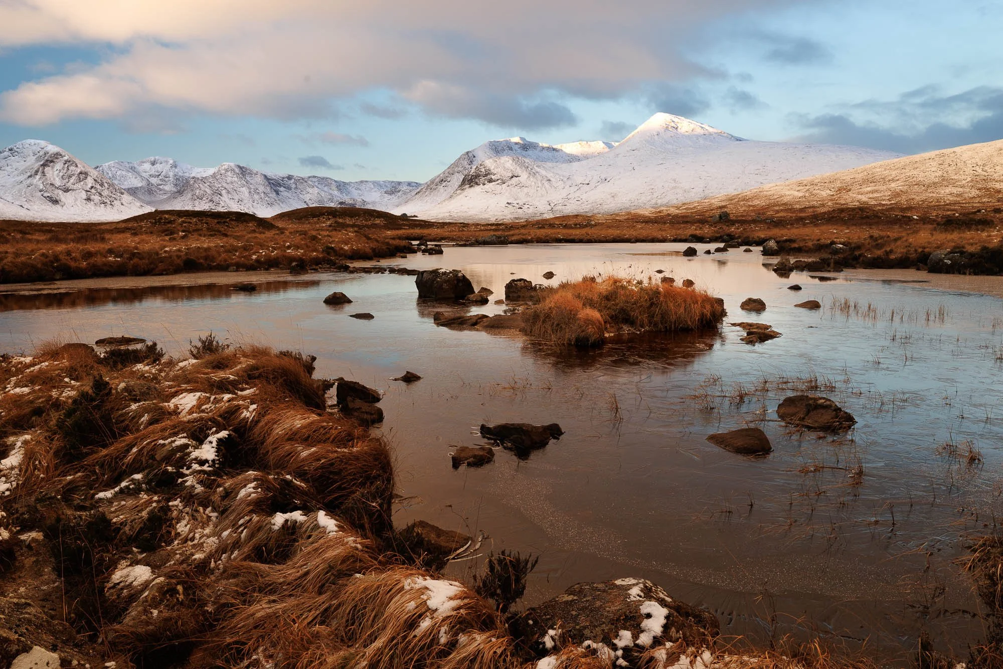 Glencoe, Scotland, snow capped mountains in the background with a river flowing through a grassy landscape in the foreground.