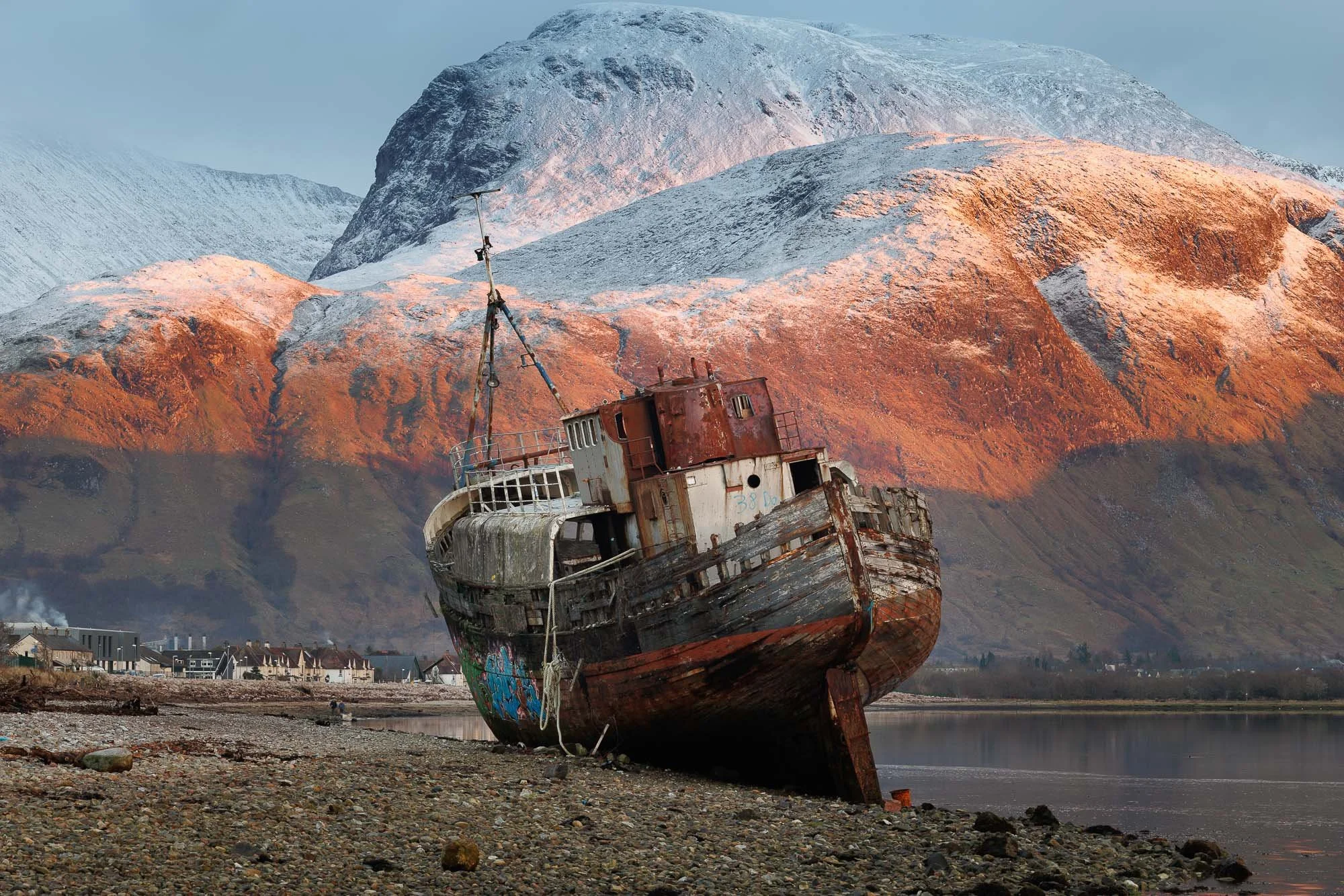 An abandoned, rusted shipwreck on a pebbled shore with Ben Nevis, Scotland, in the background.