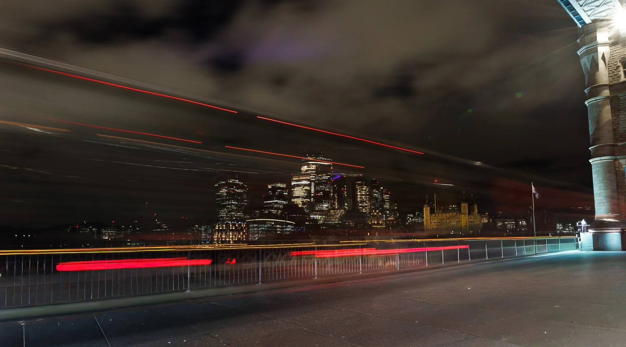 Nighttime cityscape of a downtown area viewed from a bridge with moving lights creating light trails, and a historic stone building on the right side.