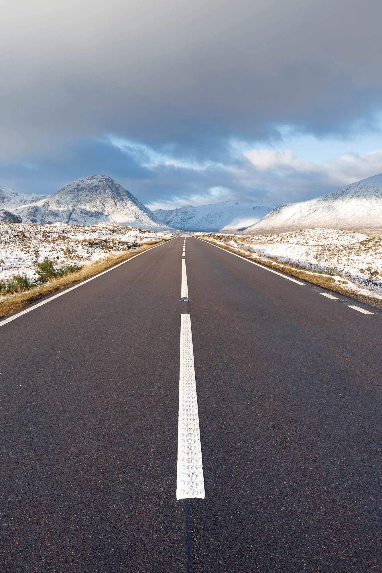 Glencoe, Scotland,  straight road through snowy mountains and cloudy sky in the distance.