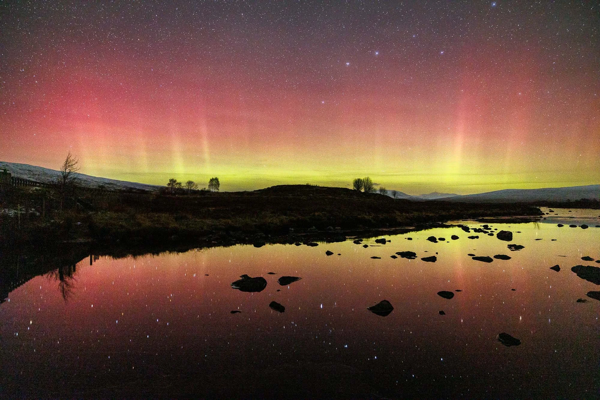 Glencoe, Scotland, Northern lights or aurora borealis over a landscape with a river and scattered rocks, with a starry sky above.