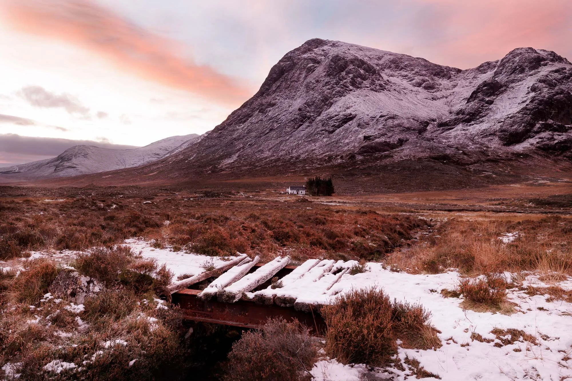 Glencoe, Scotland, snow covered bridge in a grassy field with mountains in the background at sunset.