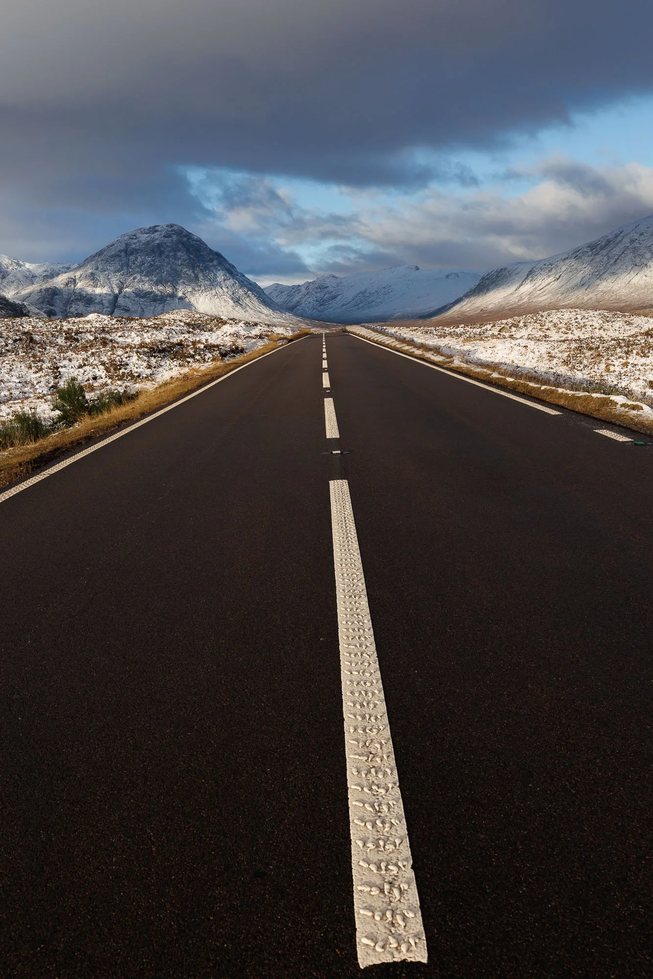 Glencoe, Scotland,  straight black asphalt road with a white dashed center line extending into the distance, surrounded by snow-covered rocky terrain and mountains under a cloudy sky.