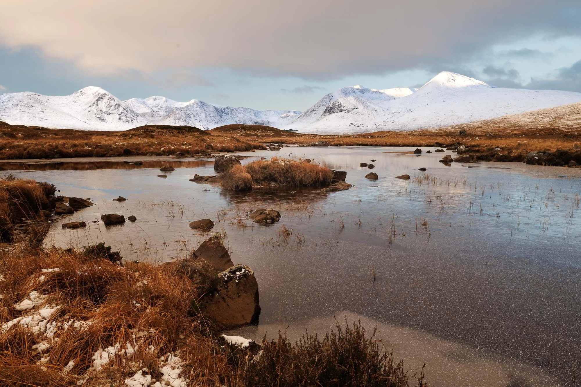 Glencoe, Scotland, snow capped mountains in the background, a calm river with rocks and tufts of grass in the foreground, and a cloudy sky overhead.