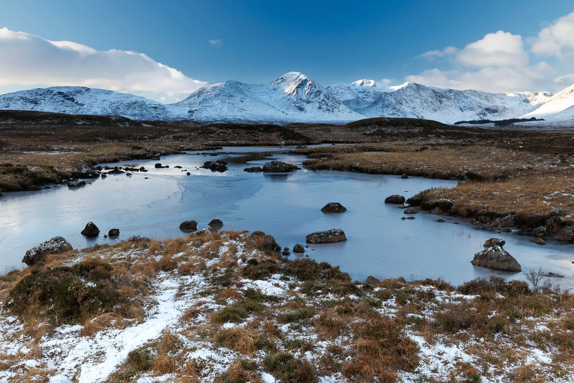 Snow-capped mountains in the background with a partly cloudy sky, a meandering icy river in the foreground, and dry grass with patches of snow on the ground.