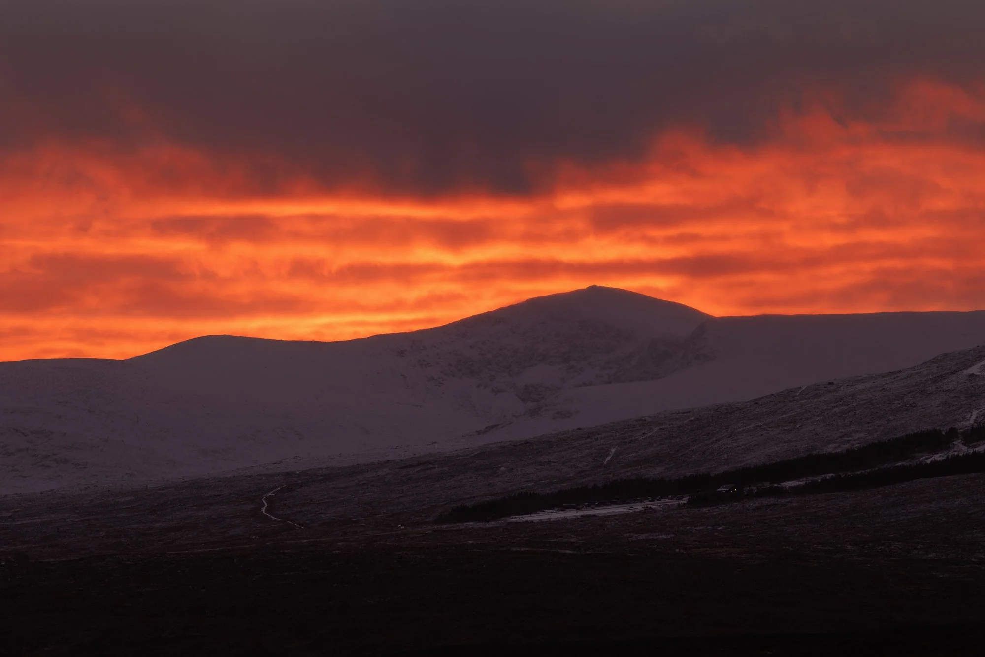  Glencoe, Scotland,  landscape at sunrise with a dark sky and orange glow, snow-covered mountain peaks, and a dark foreground.
