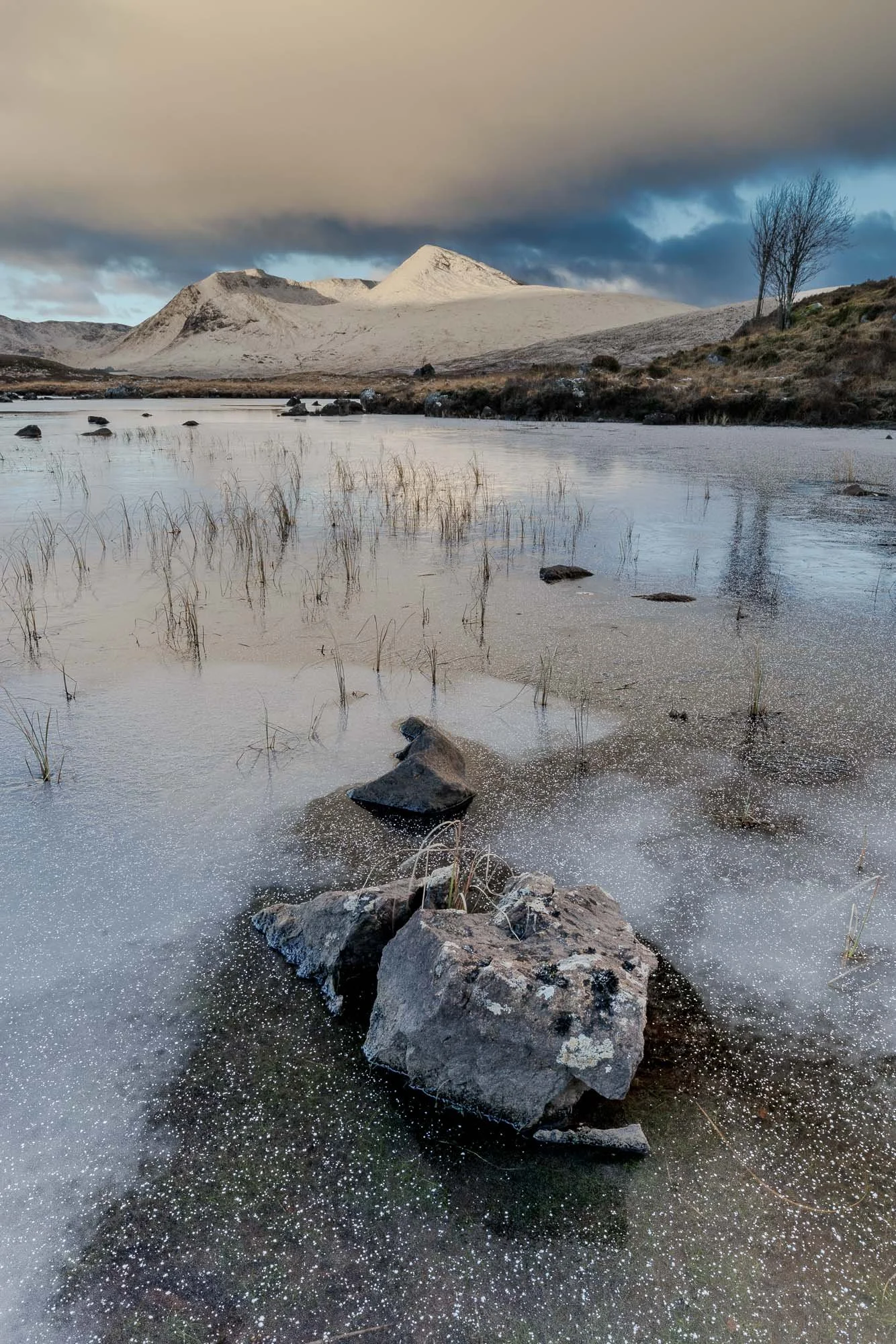 Glencoe, Scotland, snow-capped mountain in the background with dark clouds above, a partially frozen lake in the foreground with rocks and sparse grass, and a few leafless trees on the right.