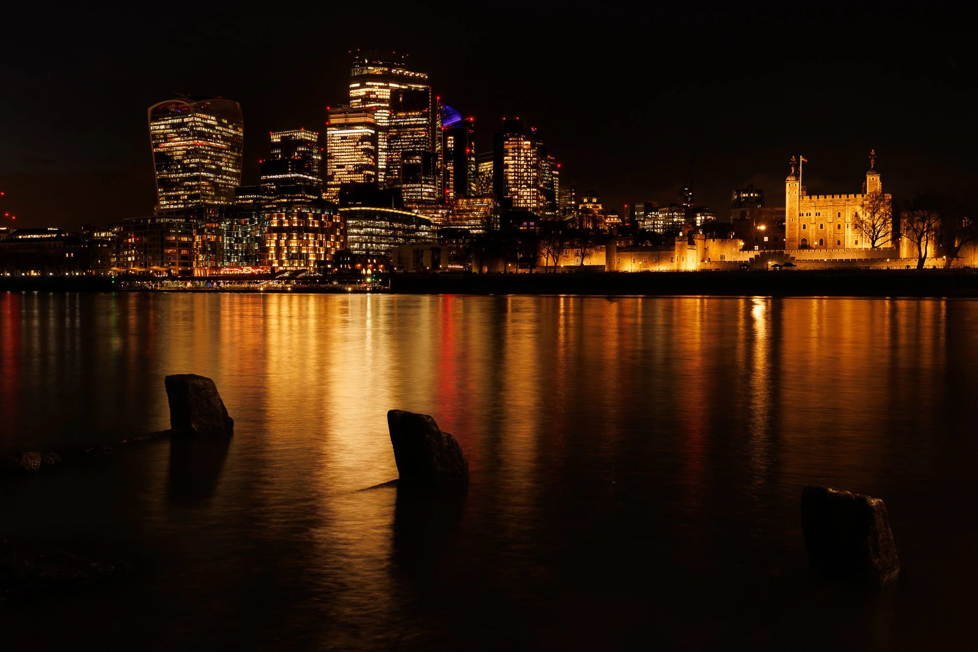 Nighttime cityscape of London with illuminated buildings reflecting on the river, featuring modern skyscrapers and historic castle walls.