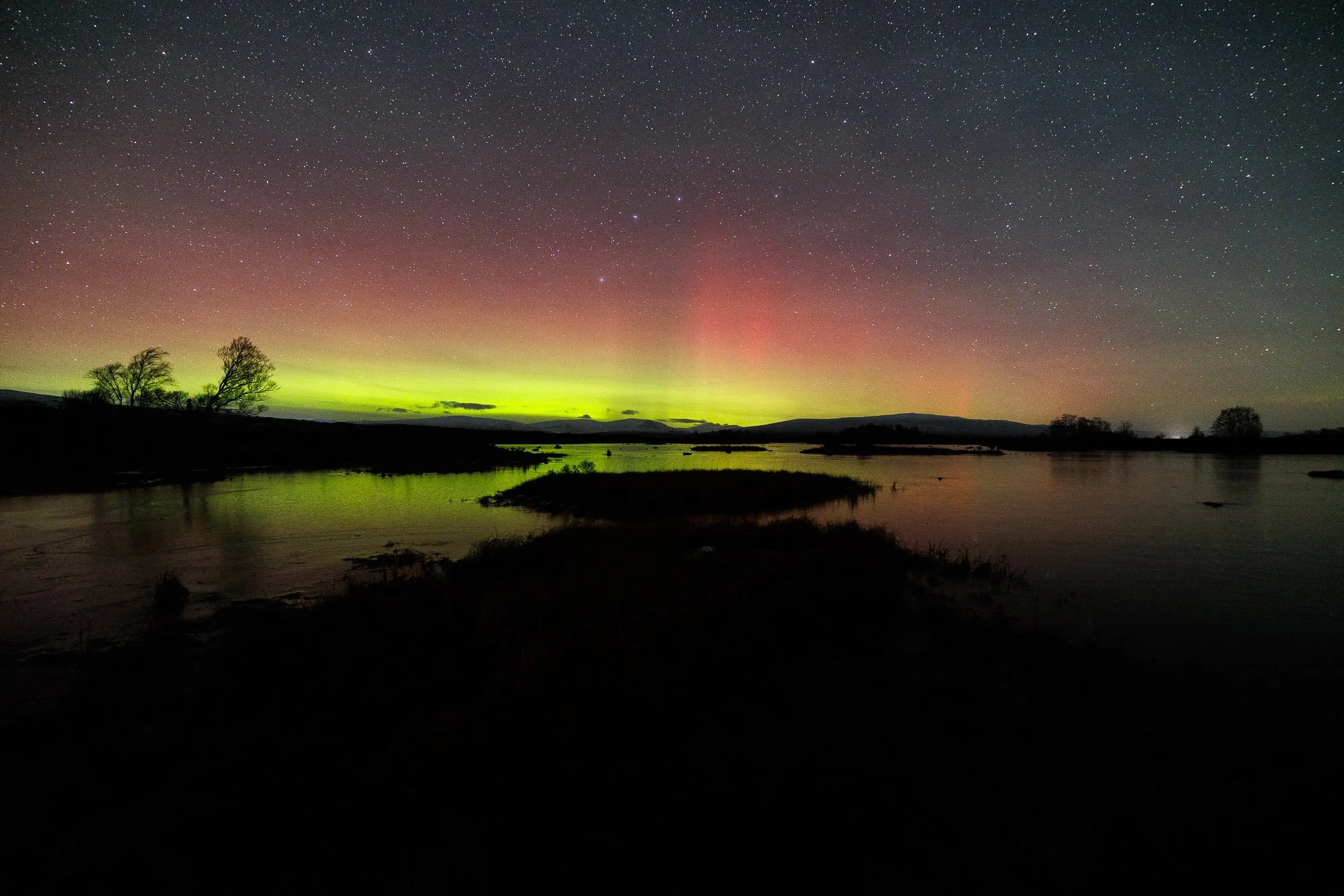 Glencoe, Scotland,  Lights over a river at night with stars in the sky and silhouetted trees.