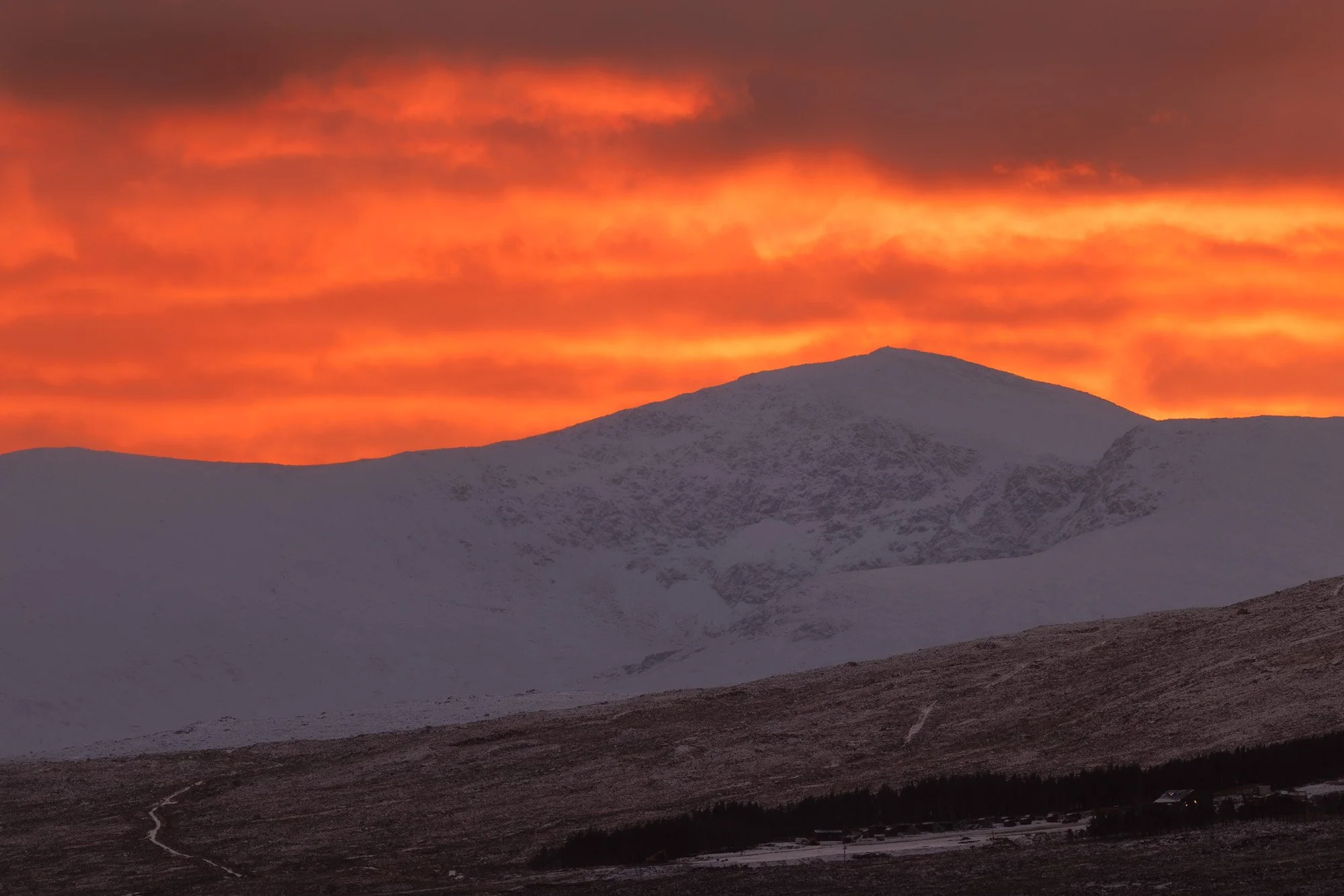 Glencoe, Scotland, snow covered mountain range at sunset with orange and pink clouds in the sky.