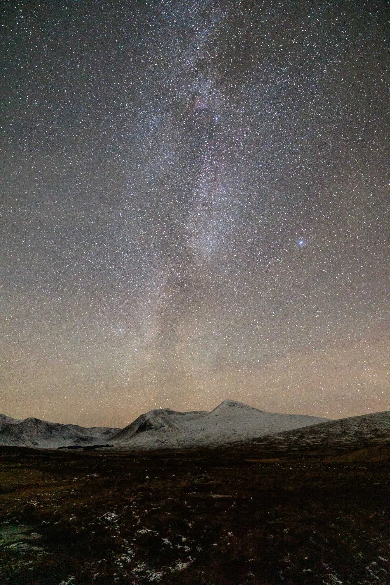 Glencoe, Scotland Night sky filled with stars and the Milky Way galaxy above snow-capped mountains and a dark landscape.