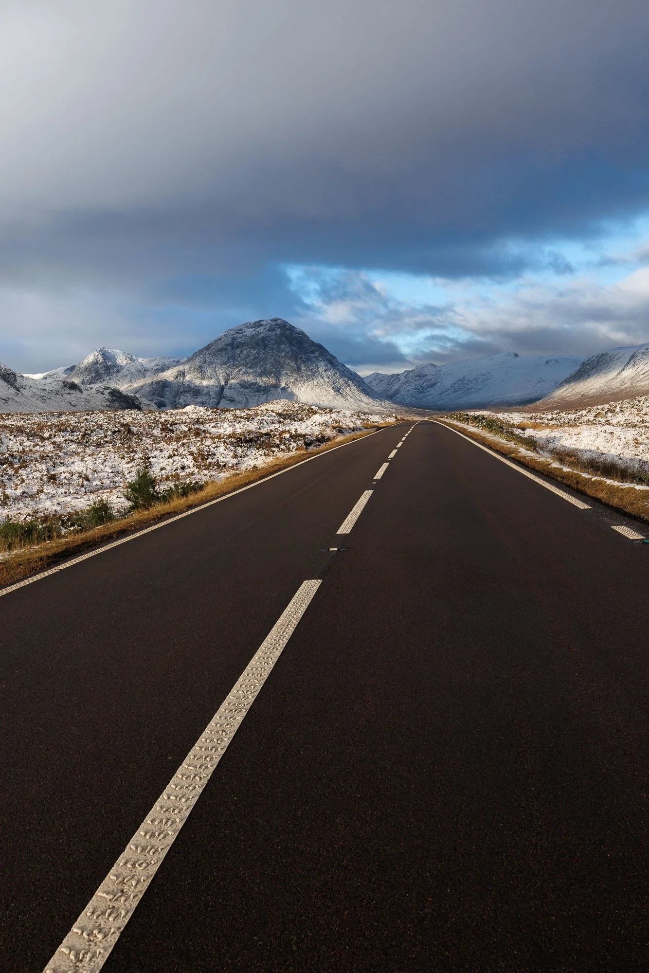 Glencoe, Scotland,  straight road leading towards snow-capped mountains under a cloudy sky.