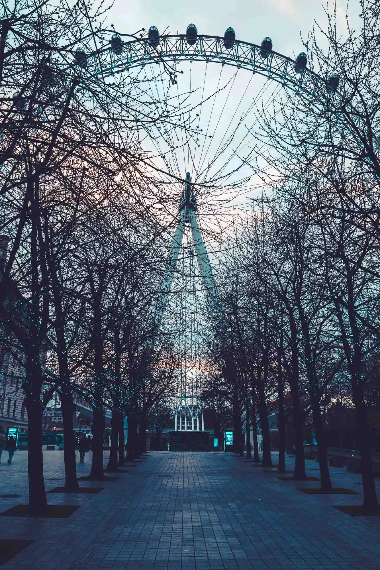 View of a large Ferris wheel viewed through leafless trees on a city street, with a cloudy sky in the background.