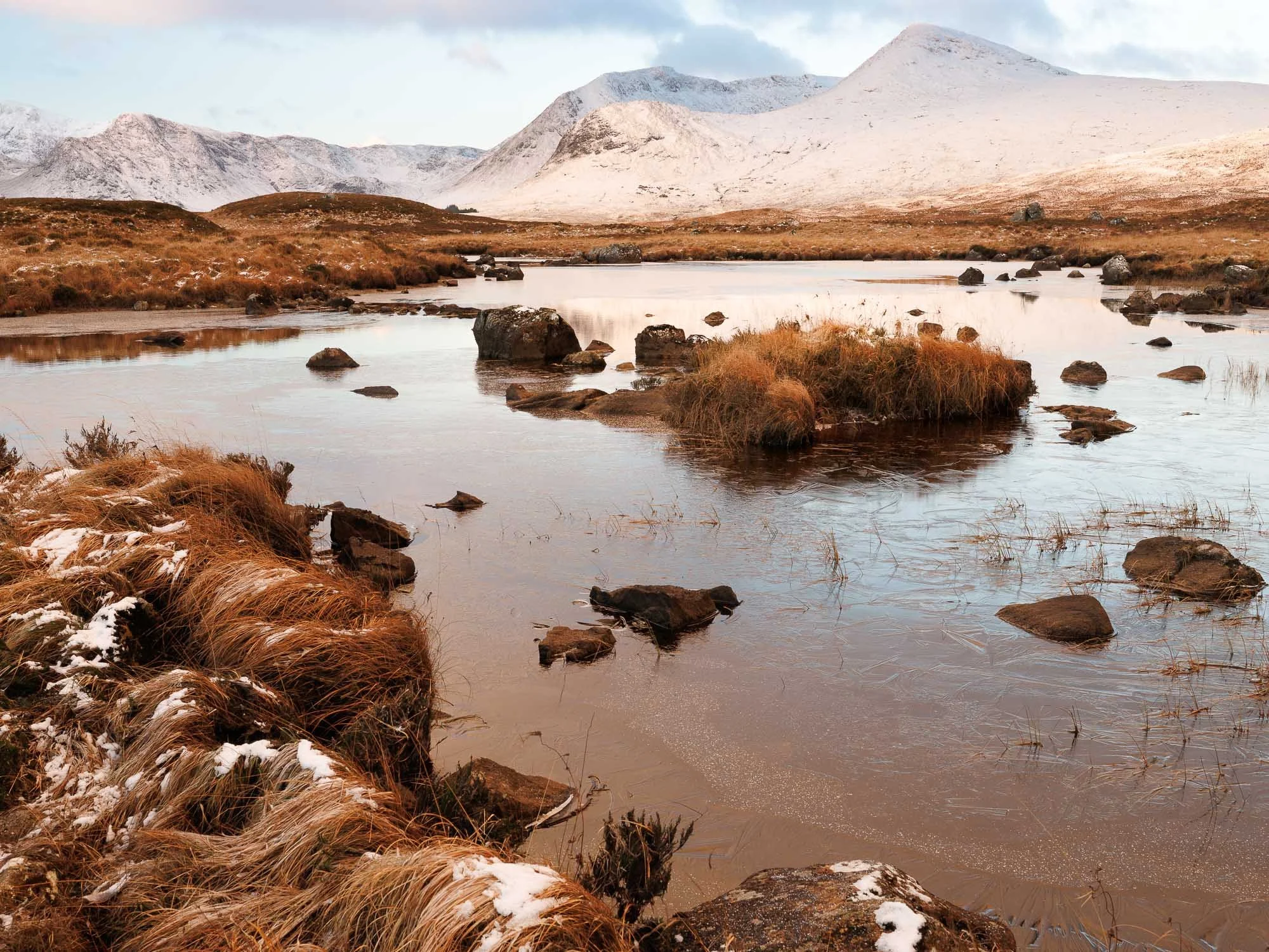 Glencoe, Scotland,  mountain lake scene with brown grasses and rocks in the foreground, calm water reflecting the sky, and snow-capped mountains in the background.