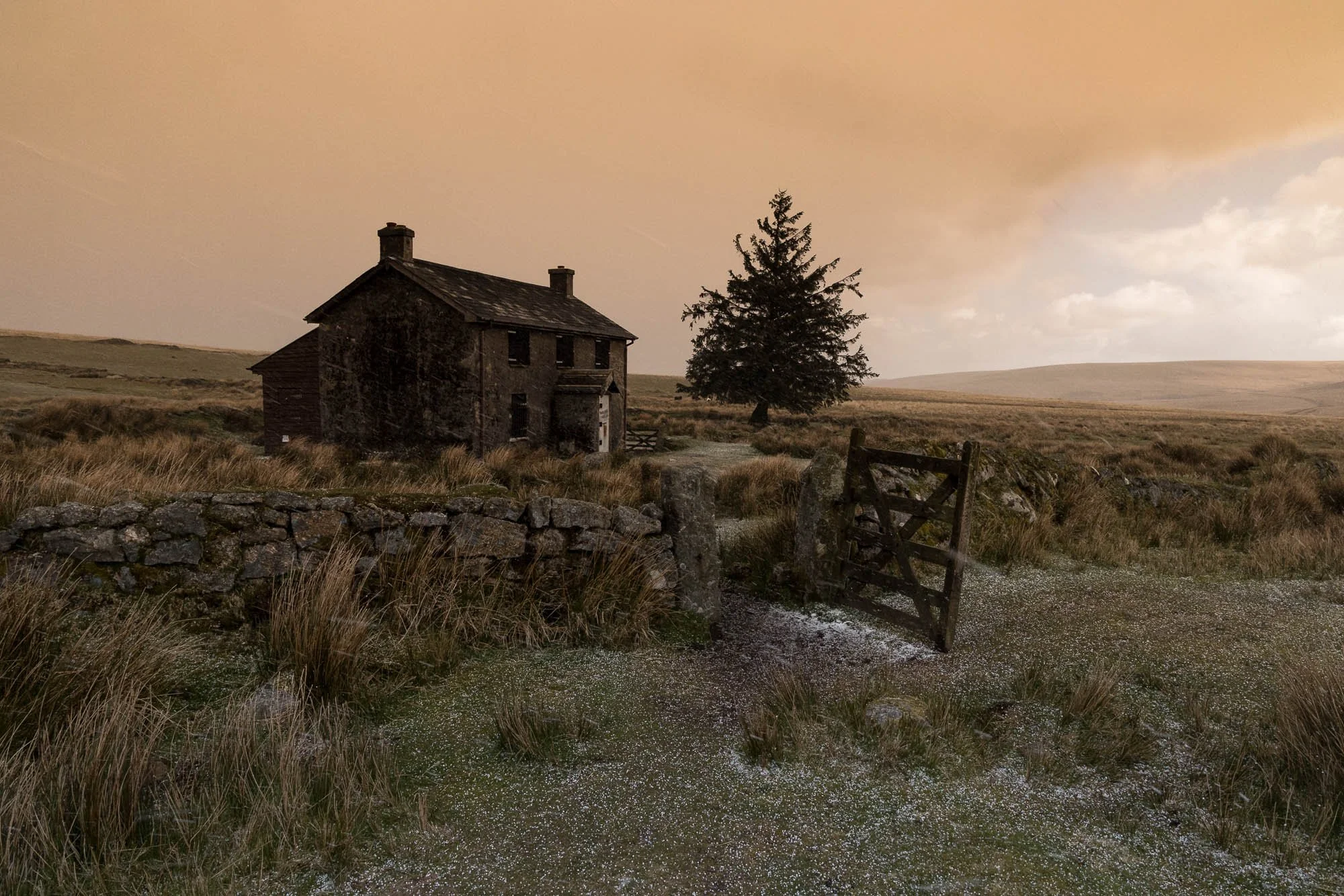An old, abandoned two-story house with a pitched roof and chimneys sits in a rural landscape. There is a large tree nearby and a stone wall with an open wooden gate in the foreground. The scene is set during a cloudy, possibly early evening, with an 