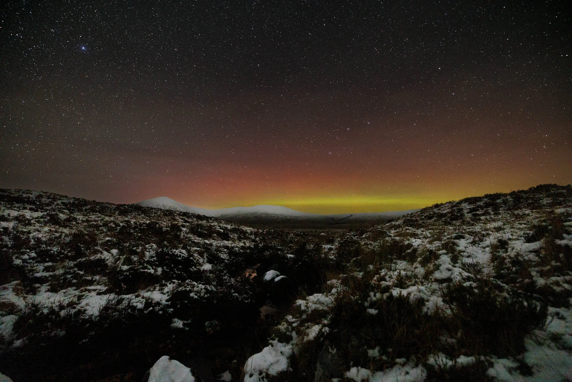 Glencoe, Scotland, landscape with snow-covered hills and a star-filled sky, showing the northern lights with green and red hues over distant mountains.